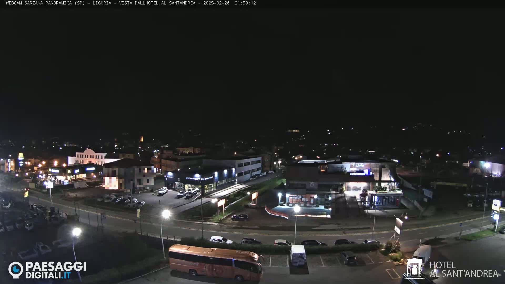 A nighttime, aerial view of a town's outskirts, showing various buildings including restaurants and a hotel, with cars parked in lots and a large bus parked near a roadway.