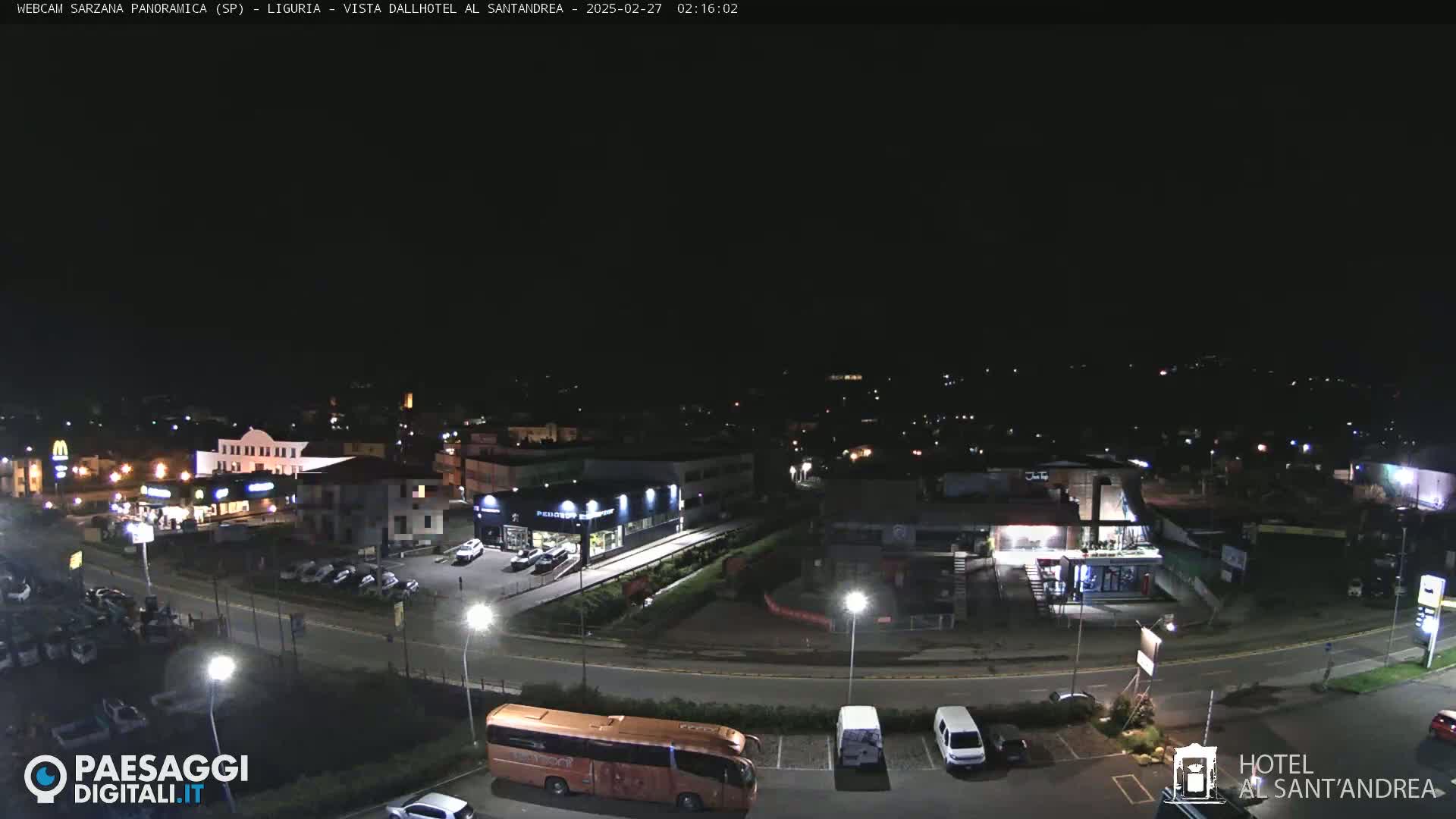 A nighttime, high-angle view of a town's outskirts, showing roads, buildings, parked cars, and a bus, under a clear, dark sky.