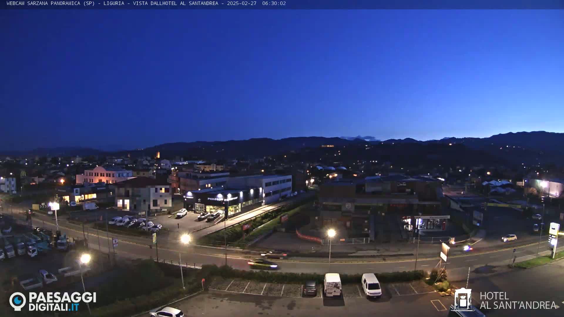 A nighttime view of a town, illuminated by streetlights, nestled against a dark mountain range under a clear, twilight sky.
