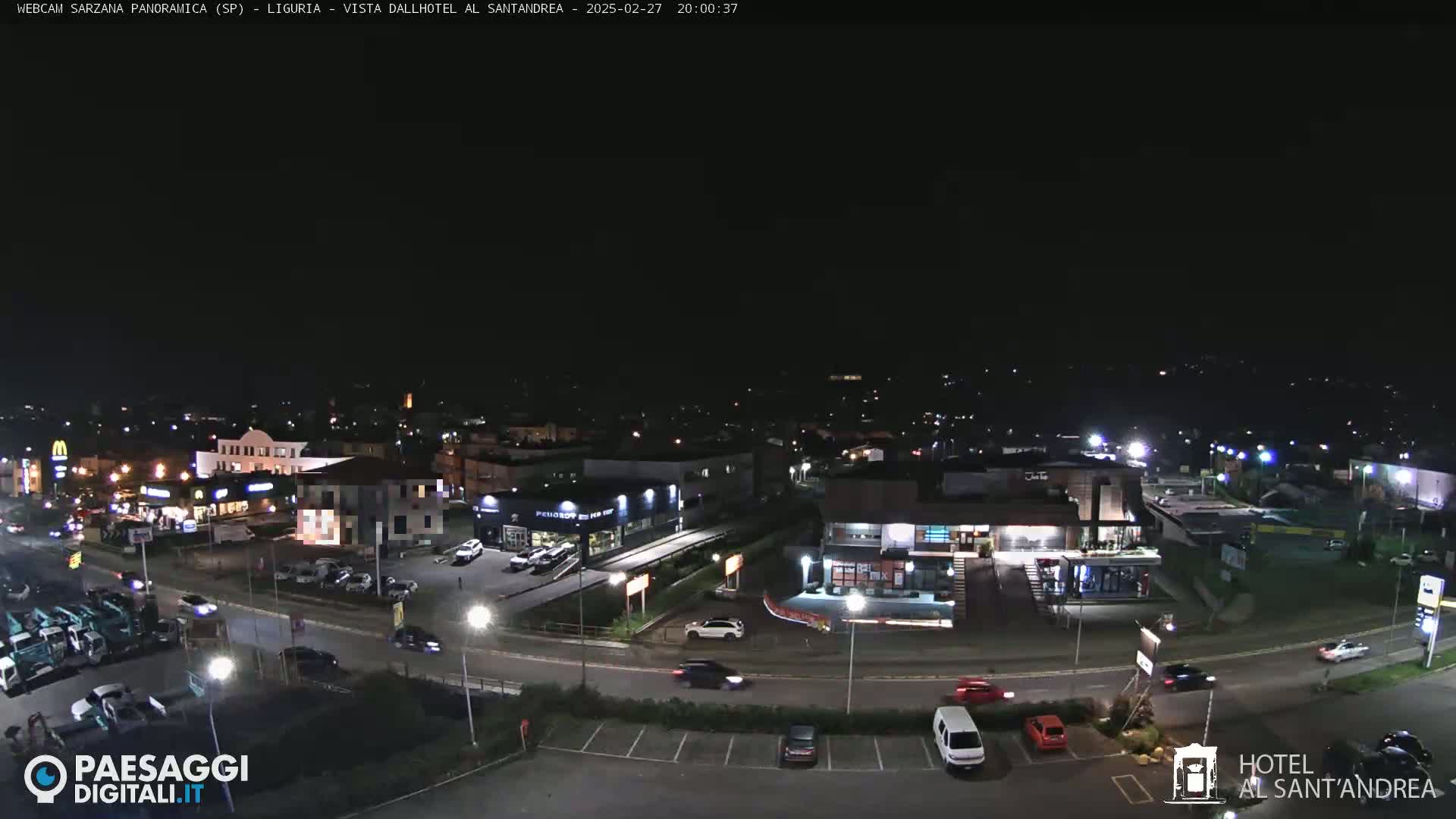 A nighttime, overhead view of a town's streets and buildings, including restaurants and a car dealership, with cars driving on the roads and parked in lots.