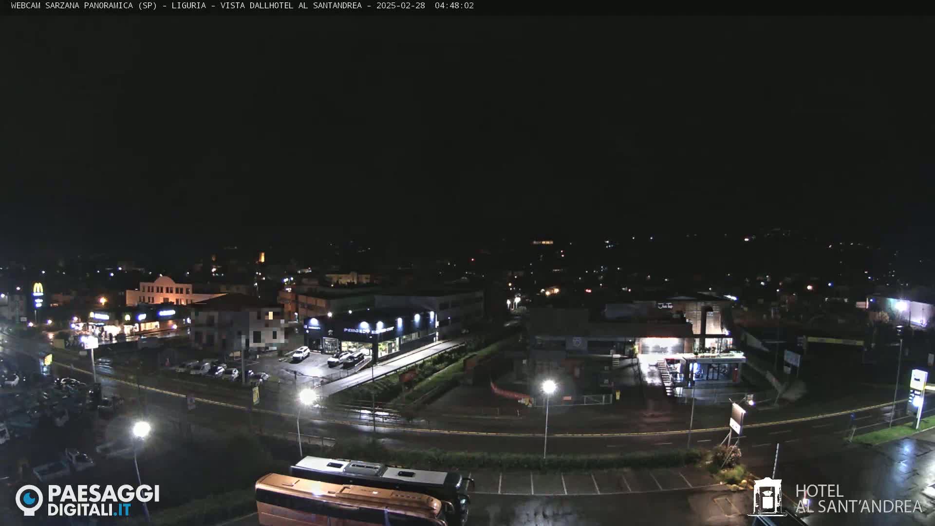 A nighttime, wet view of a town's streets and buildings, with cars parked in lots and a bus visible.