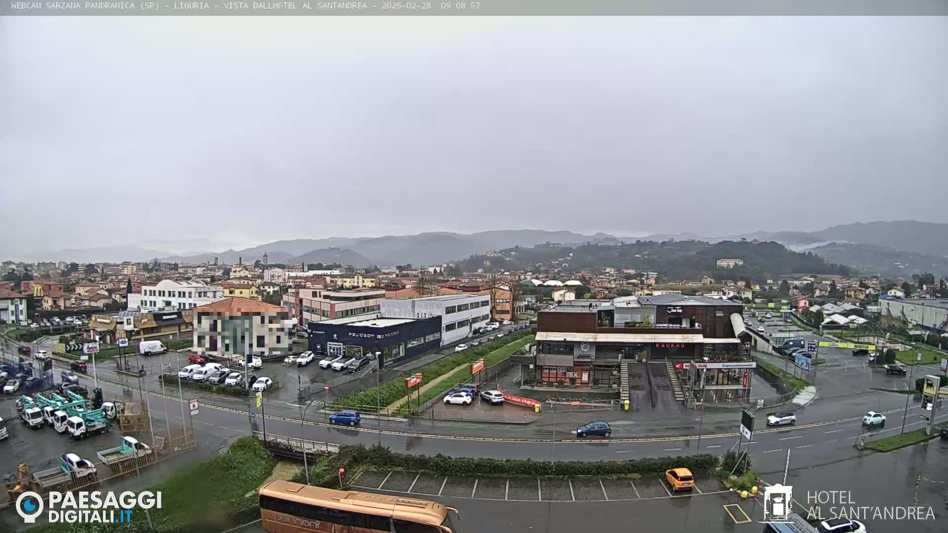 A wet, overcast cityscape featuring a road intersection with various vehicles, buildings, and a hillside in the background.