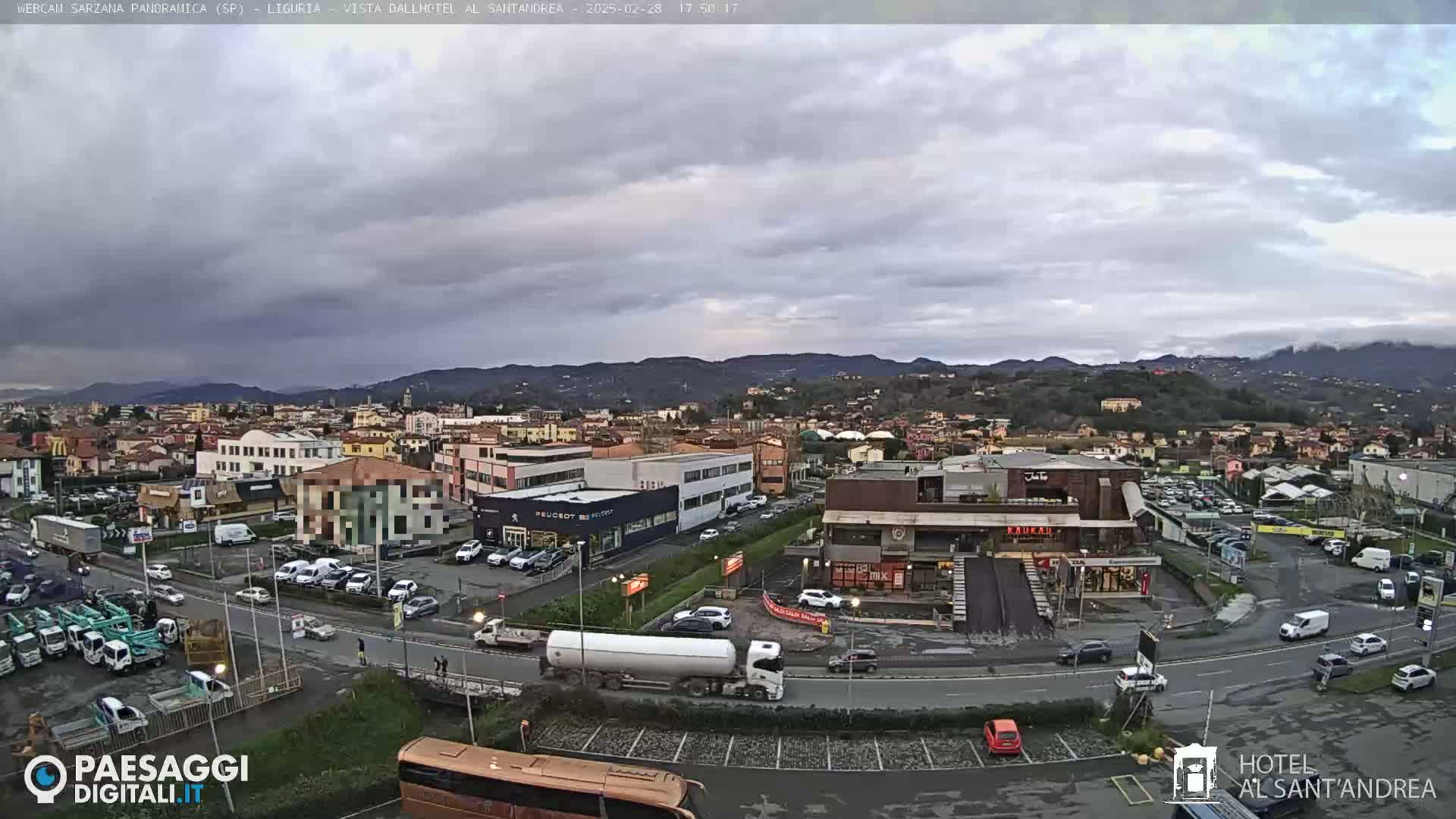 A wide shot of a town under a cloudy sky, showing buildings, roads, vehicles, and distant hills.