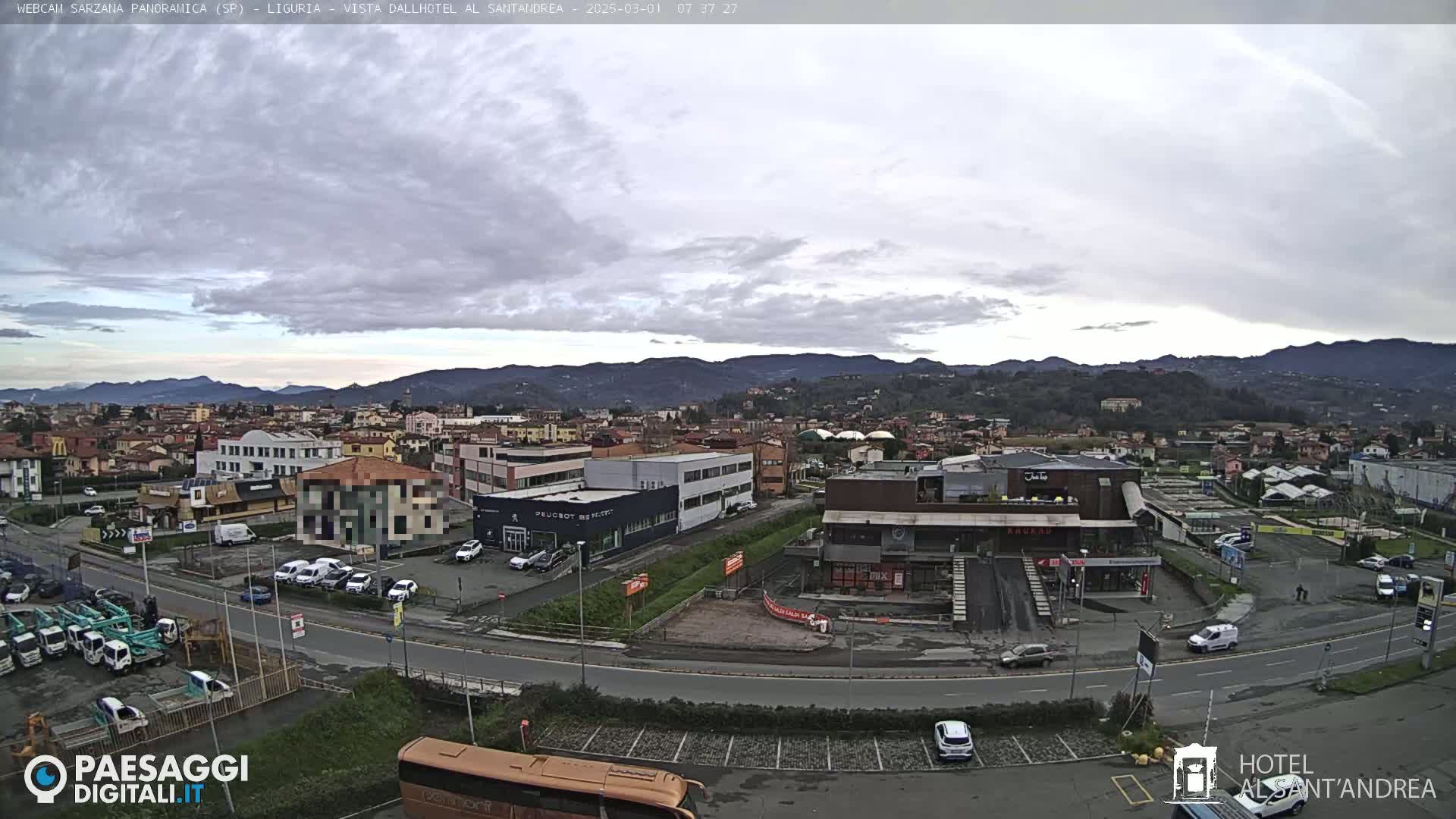 A panoramic view of a town under an overcast sky, showing buildings, roads, vehicles, and distant mountains.
