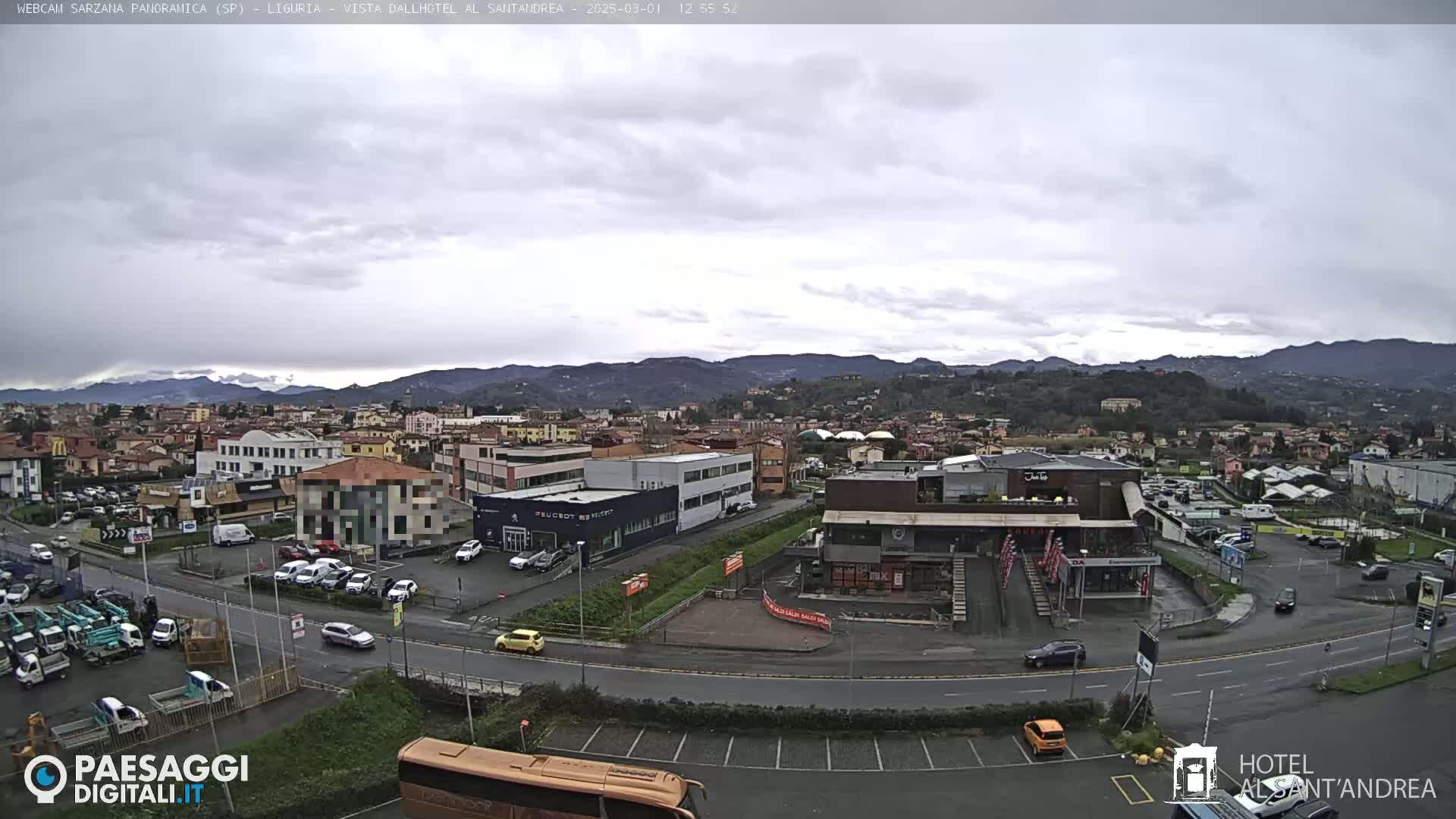 A panoramic view of a town nestled among hills under a cloudy sky shows several buildings, parking lots with cars, and a road with vehicles.