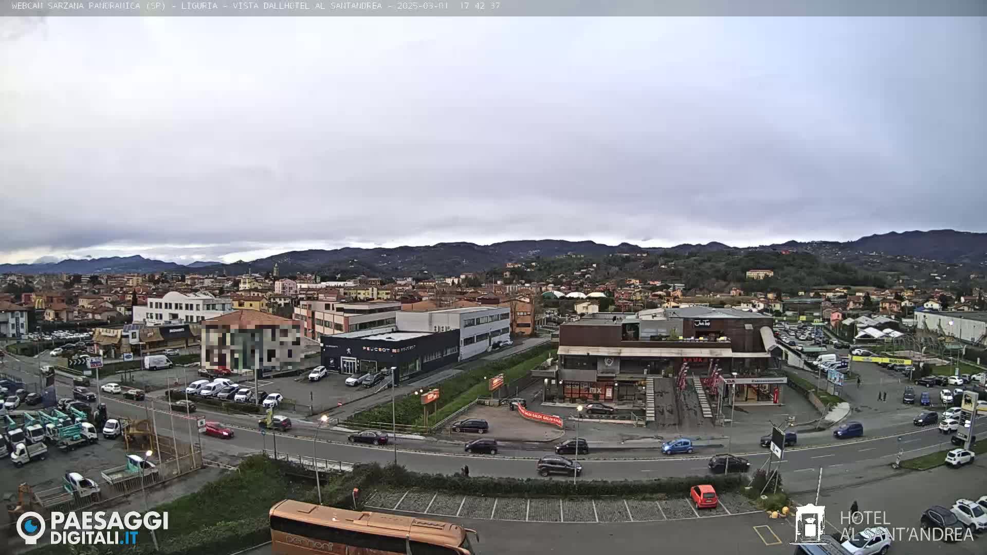 A wide, high-angle view of a town on a cloudy day, showing buildings, roads, parked cars, and a bus, with distant mountains in the background.