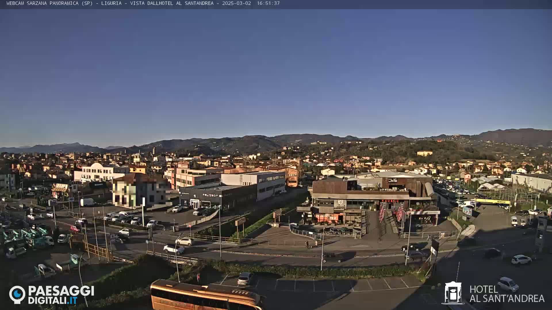 A panoramic view of a town on a clear, sunny day, showing buildings, roads, parked cars, and a bus, with hills in the background.
