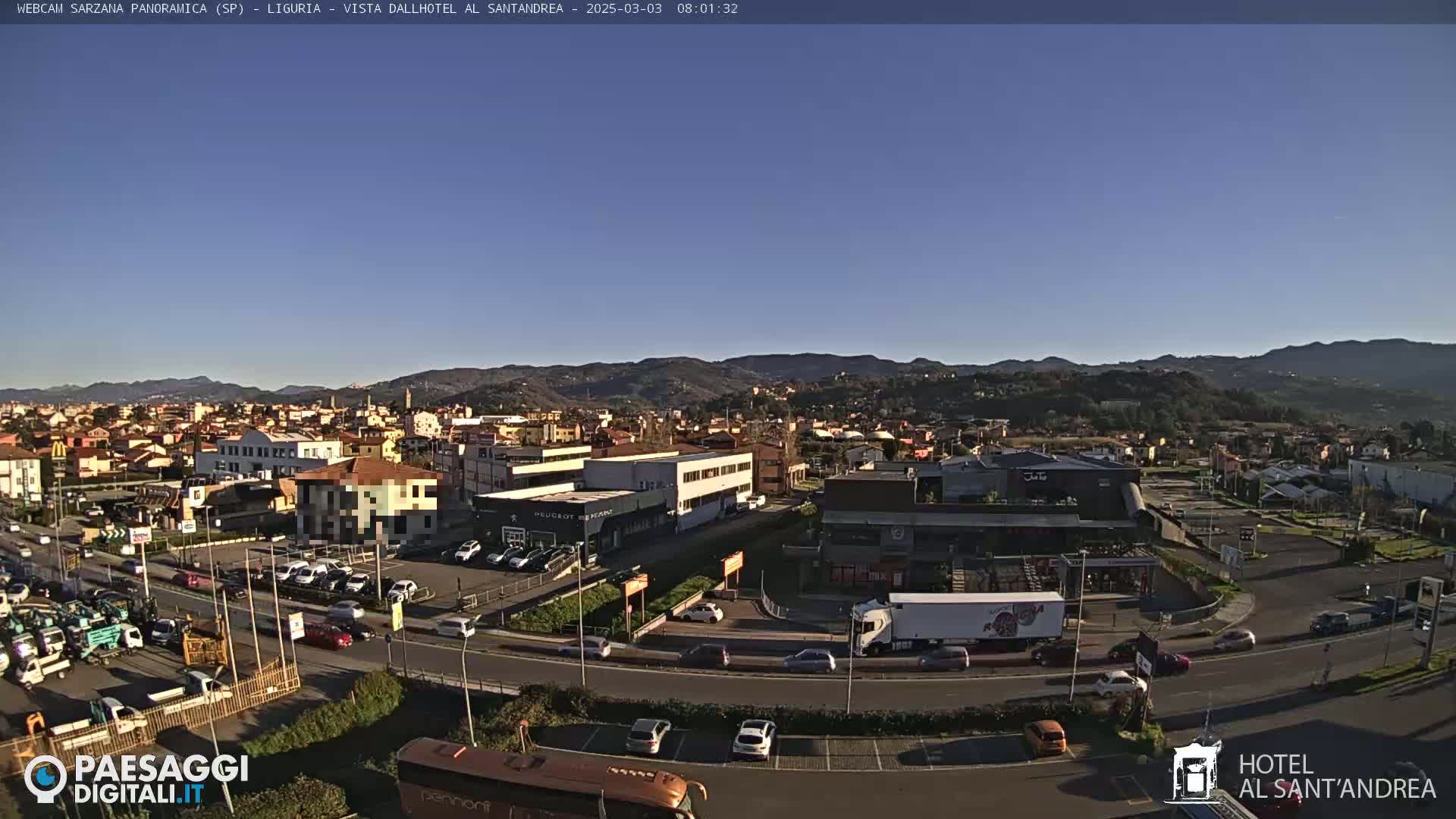 A clear, sunny day reveals a panoramic view of a town nestled among rolling hills, with cars moving along roads and a parking lot filled with vehicles.