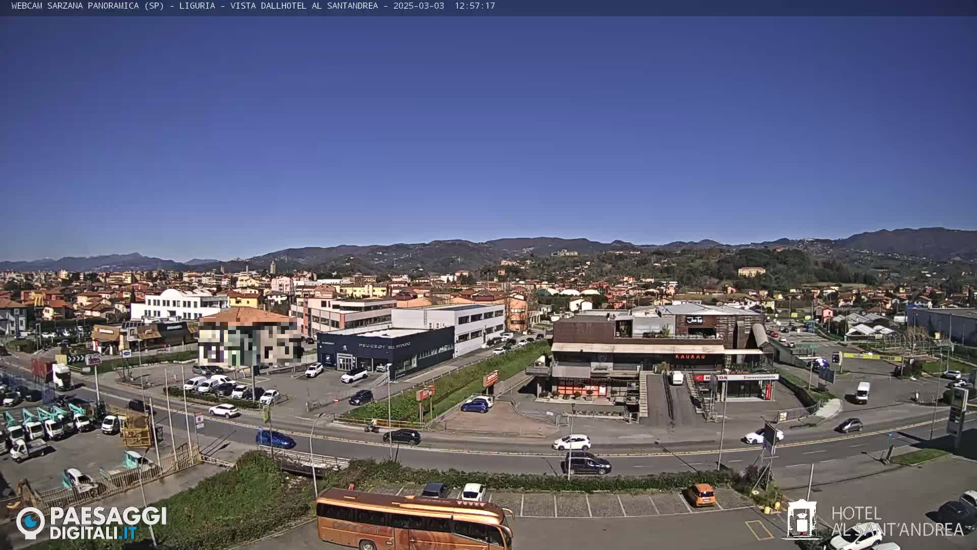 A clear, sunny day reveals a panoramic view of a town nestled amongst hills, with various buildings, cars, and a bus visible in the foreground.