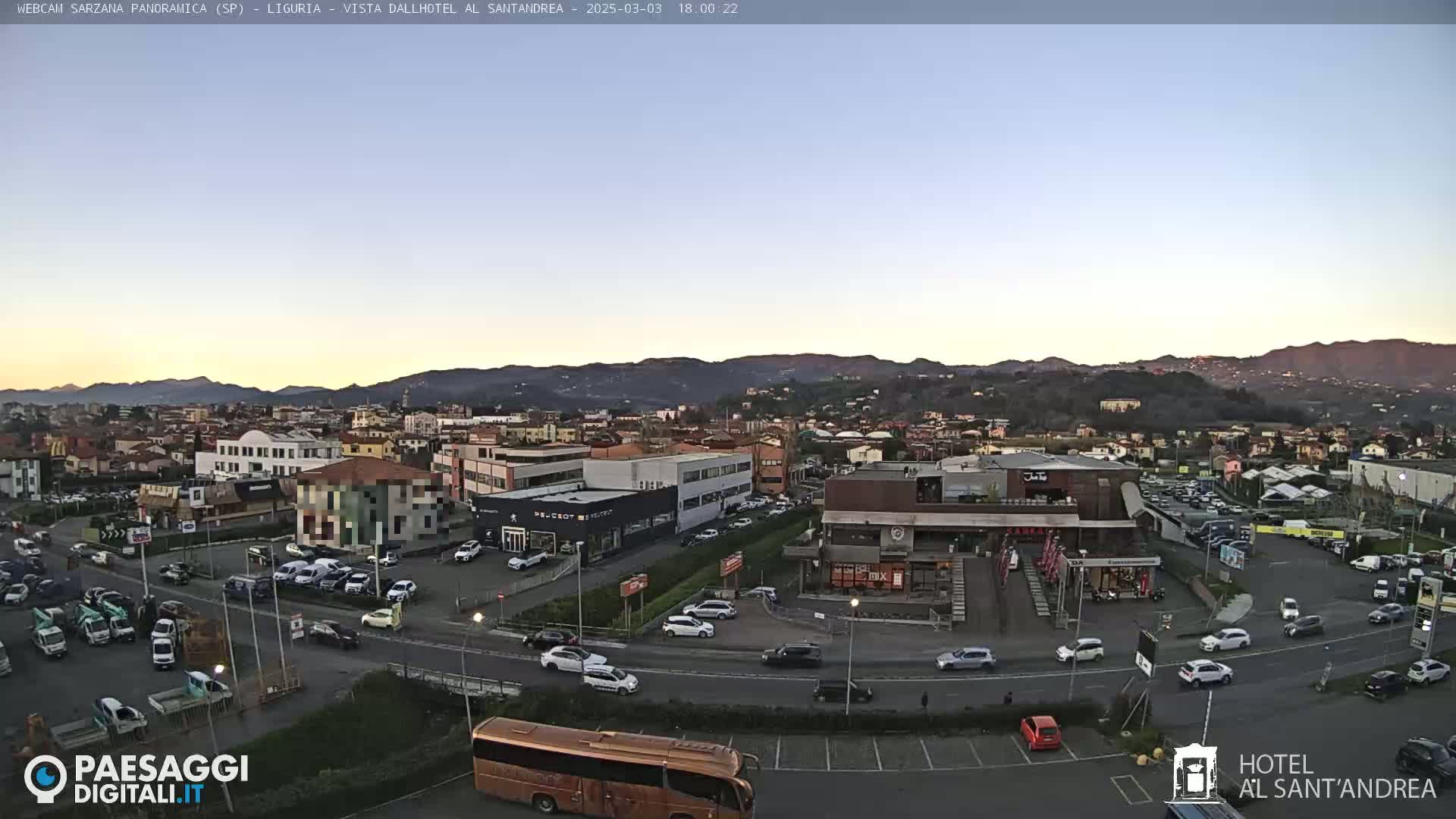 A wide aerial view of a town at dusk, showing a parking lot with numerous cars and a bus, various buildings including a hotel and restaurants, and mountains in the background under a clear sky.