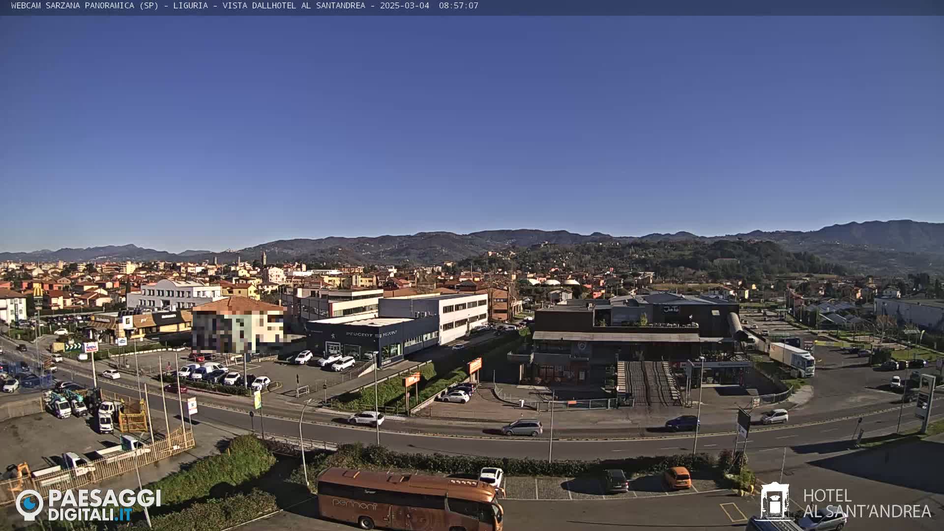 A panoramic view of a town nestled in a valley under a clear blue sky, showing buildings, roads, vehicles, and distant mountains.