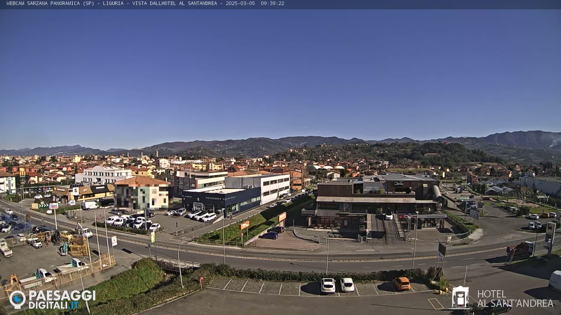 A sunny, clear day overlooking a town nestled in a valley, with mountains in the background and various buildings, roads, and parked cars visible.