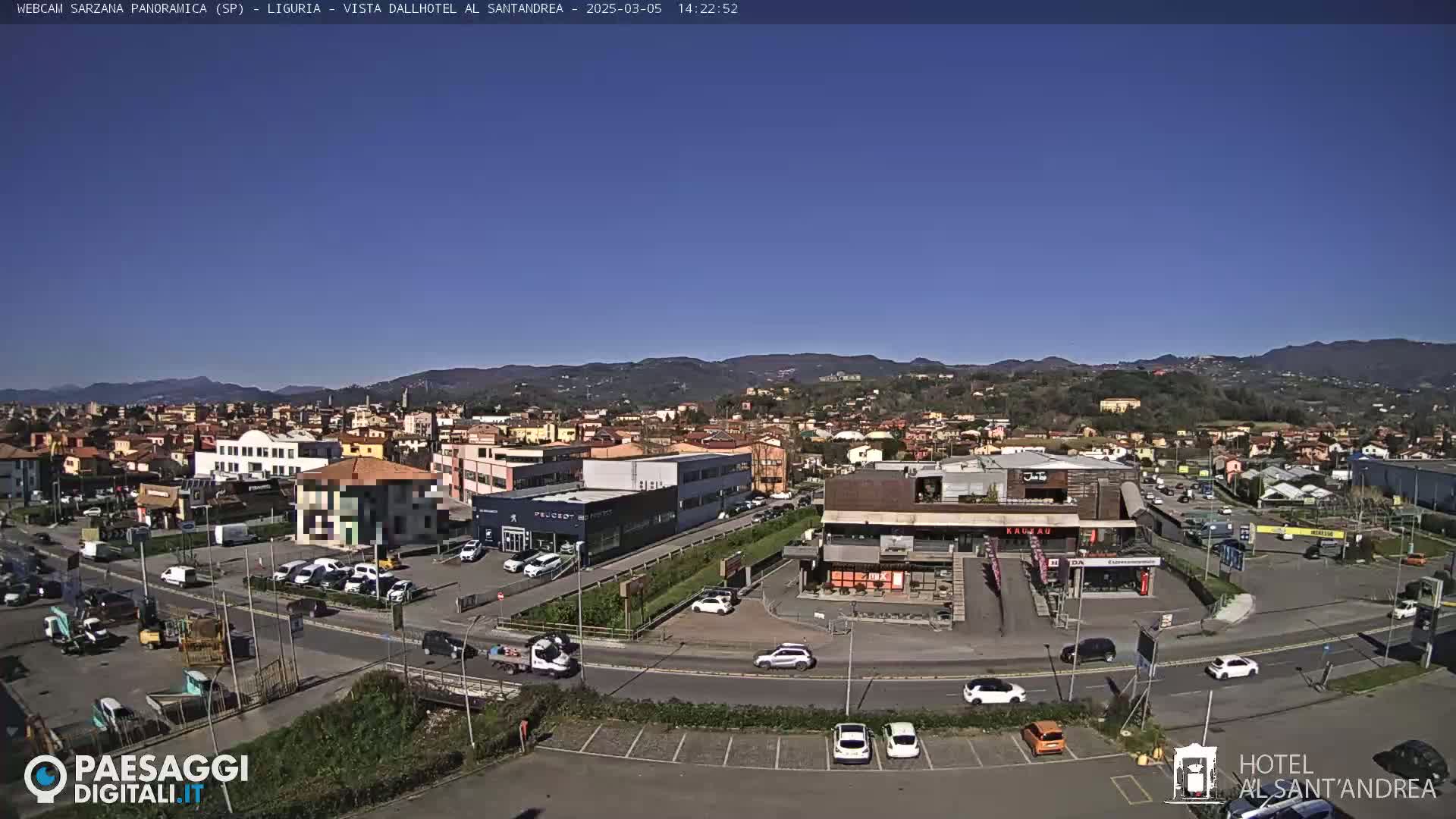 A sunny, clear day overlooks a town's cityscape, showing buildings, roads, cars in a parking lot, and distant mountains under a bright blue sky.