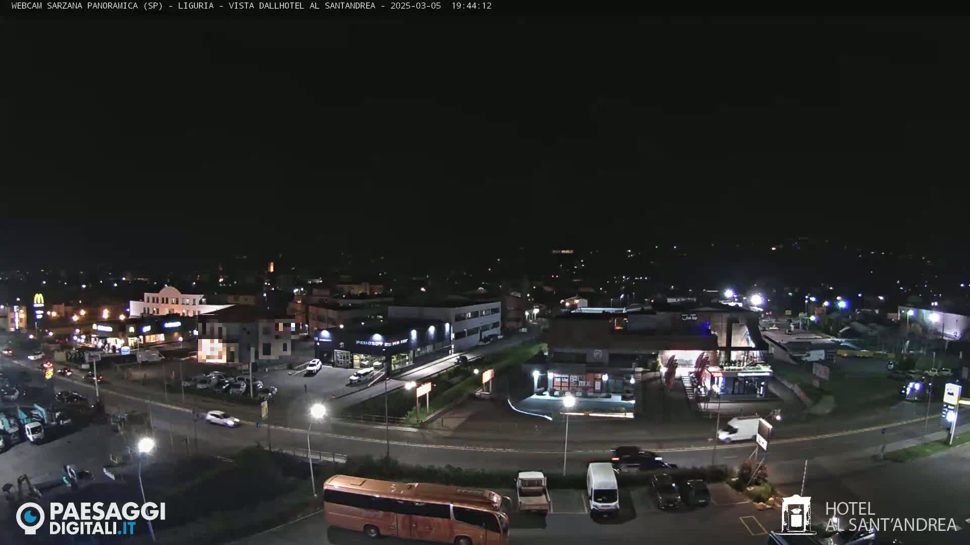 A nighttime aerial view of a town's streets and buildings, including a parking lot with cars and a bus, under a clear, dark sky.