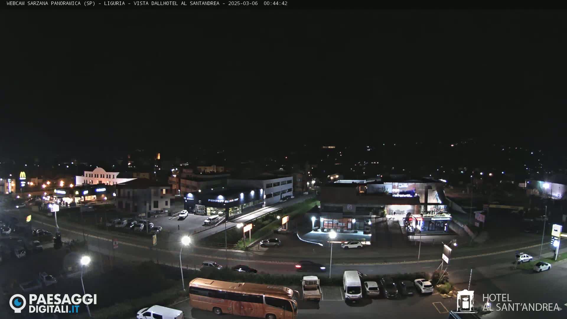 A nighttime aerial view of a town's outskirts, showing a road with parked cars and buildings, including restaurants and businesses, under a clear, dark sky.