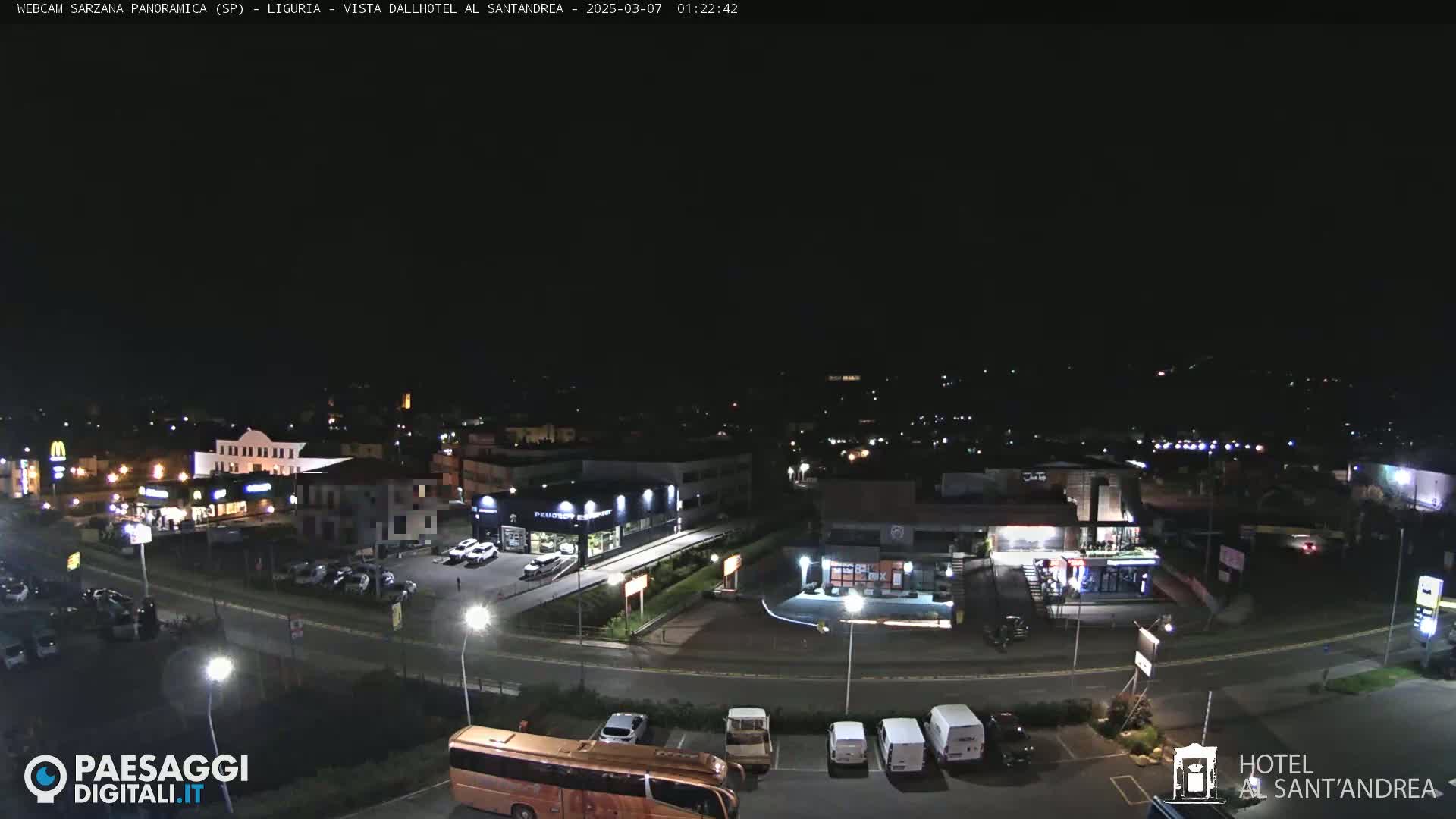 A nighttime, high-angle view of a town's outskirts, showing roadways, buildings with illuminated signs, parked cars, and a bus.