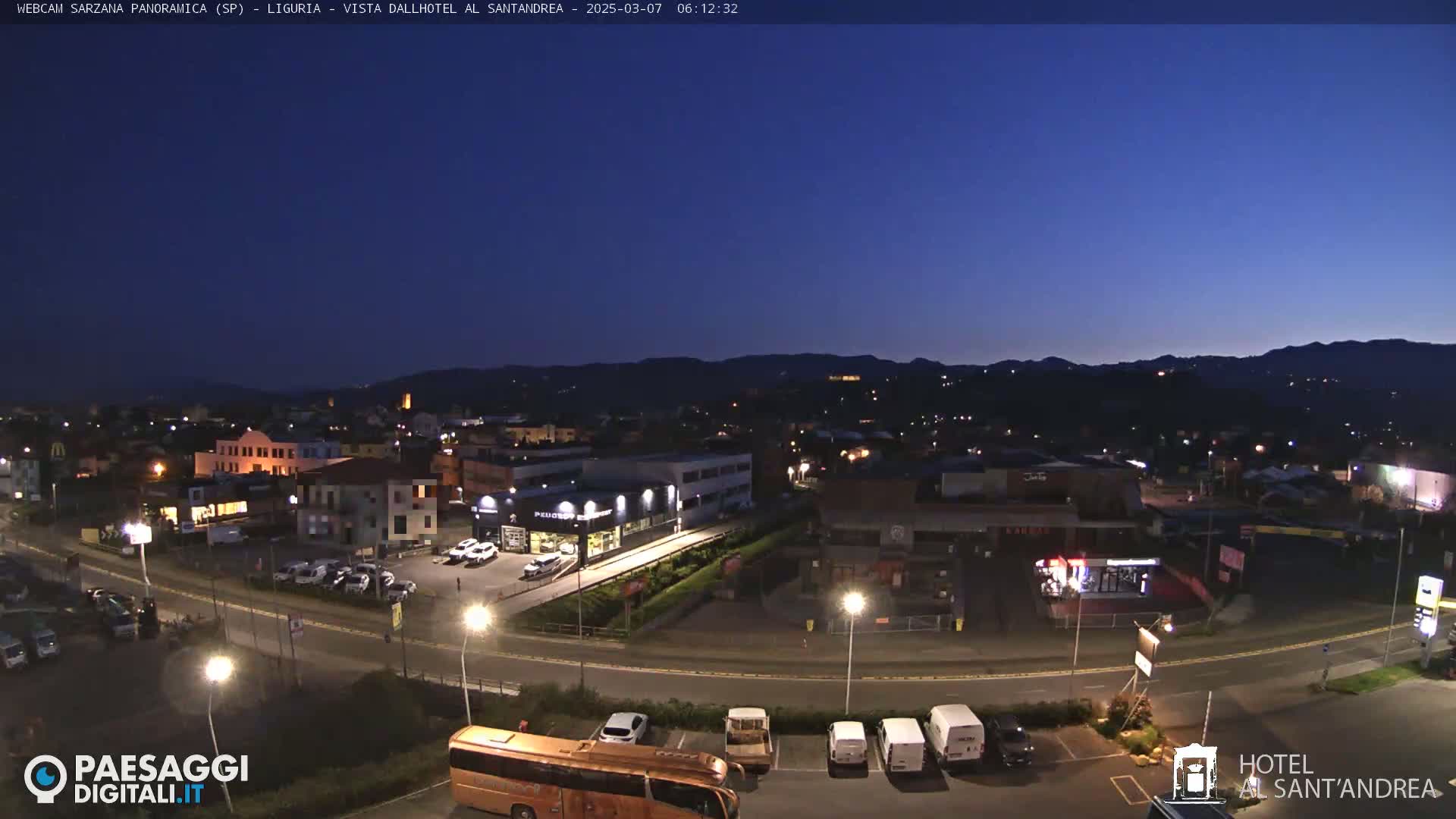 A nighttime, panoramic view of a town illuminated by streetlights, with hills visible in the background under a dark blue, twilight sky.