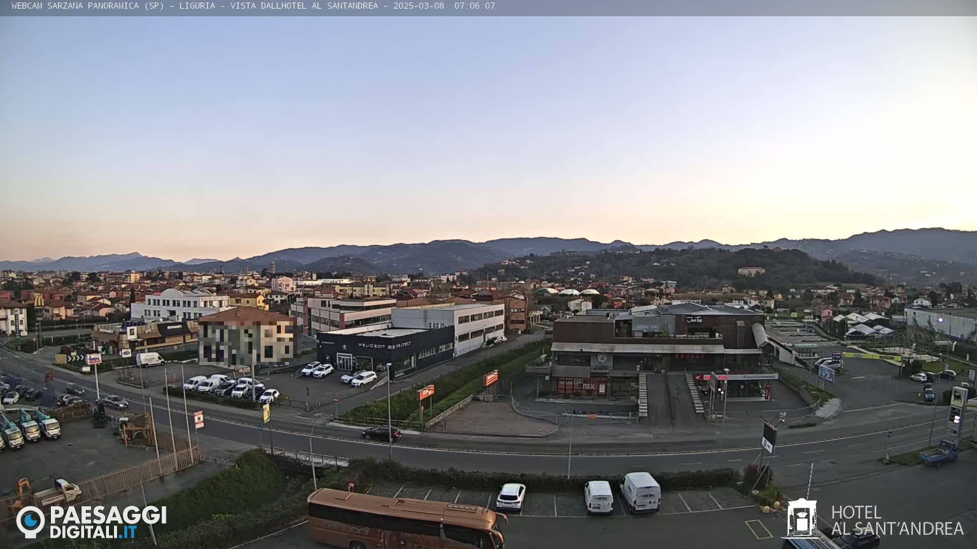 A panoramic view of a town nestled in a valley, under a clear, pale-blue twilight sky, shows buildings, parking lots, and a bus.