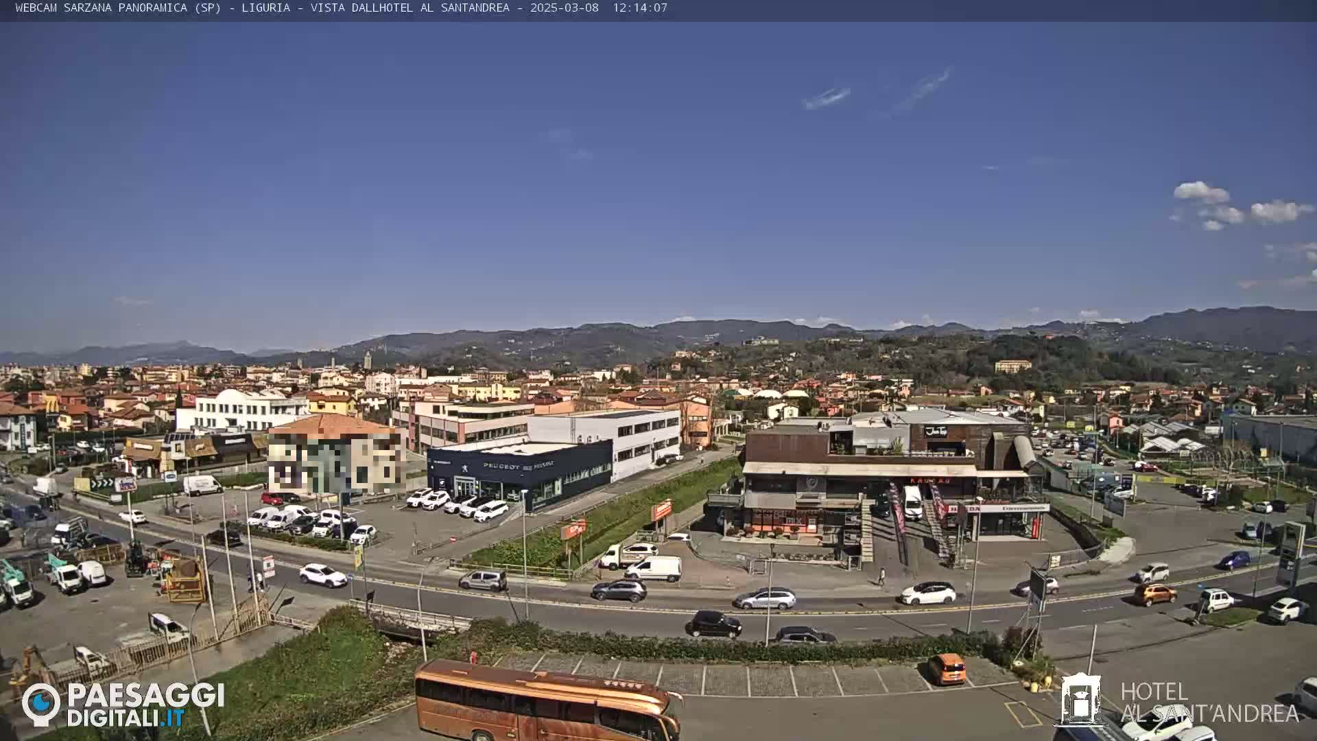 A sunny, high-angle view of a town situated in a valley, with mountains in the background and numerous cars parked in lots and driving on roads.