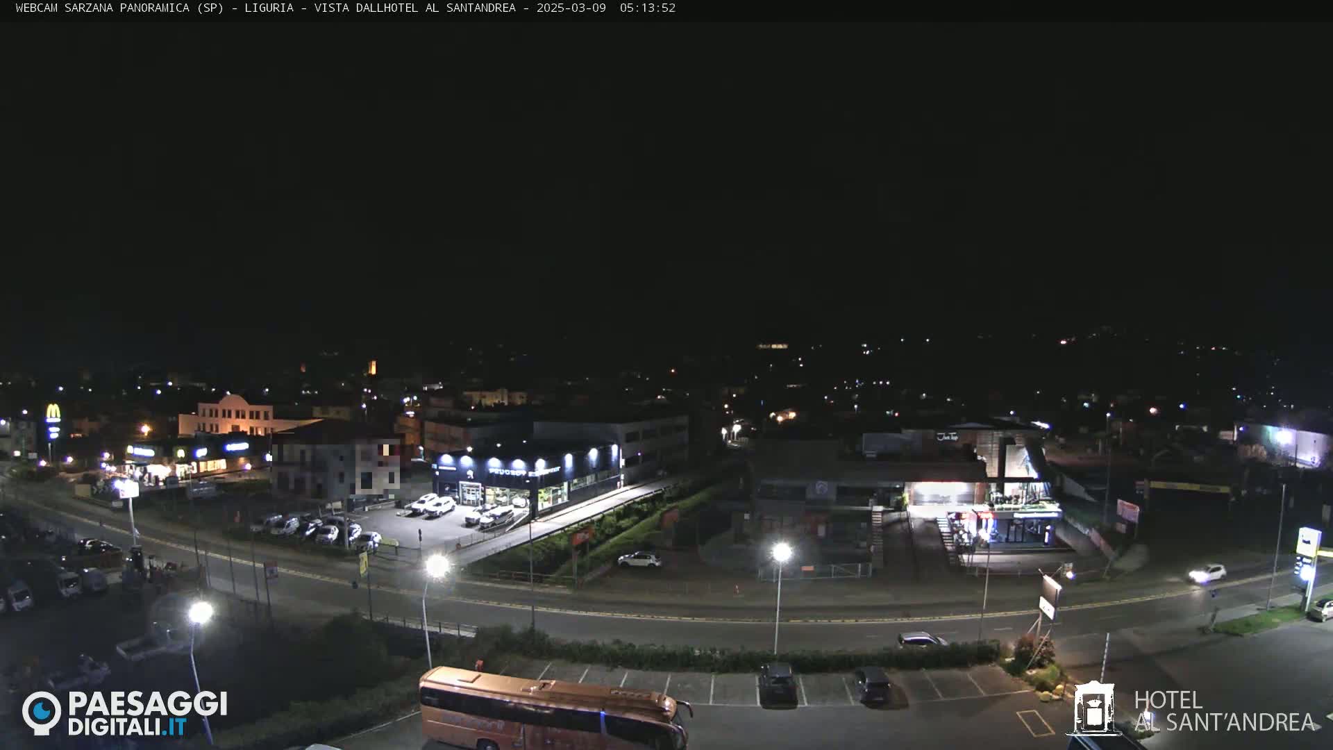 A nighttime, overhead view of a town's outskirts, featuring roads, buildings, parked cars, and a large bus.