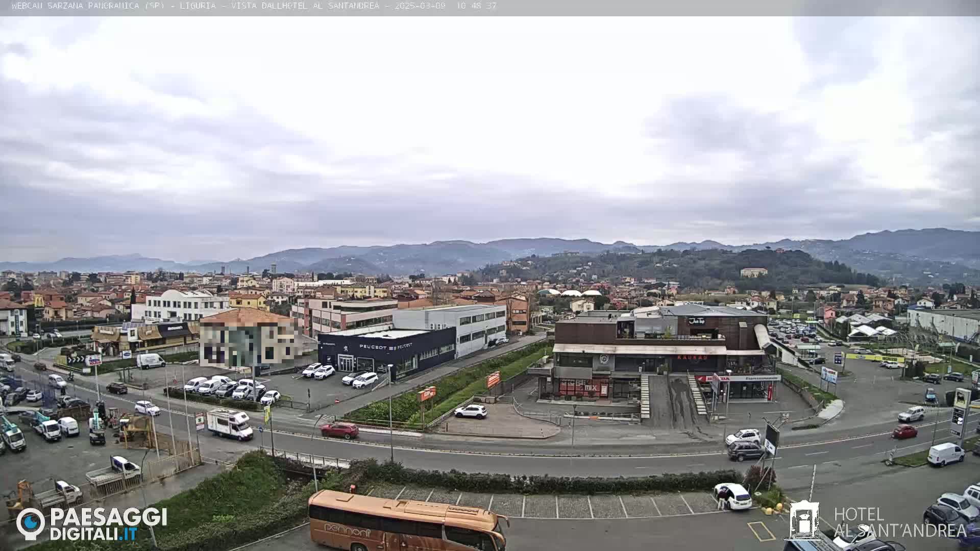 A mostly cloudy day reveals a sprawling town nestled among rolling hills, with a busy roadway featuring cars, buses, and a parking lot visible in the foreground.