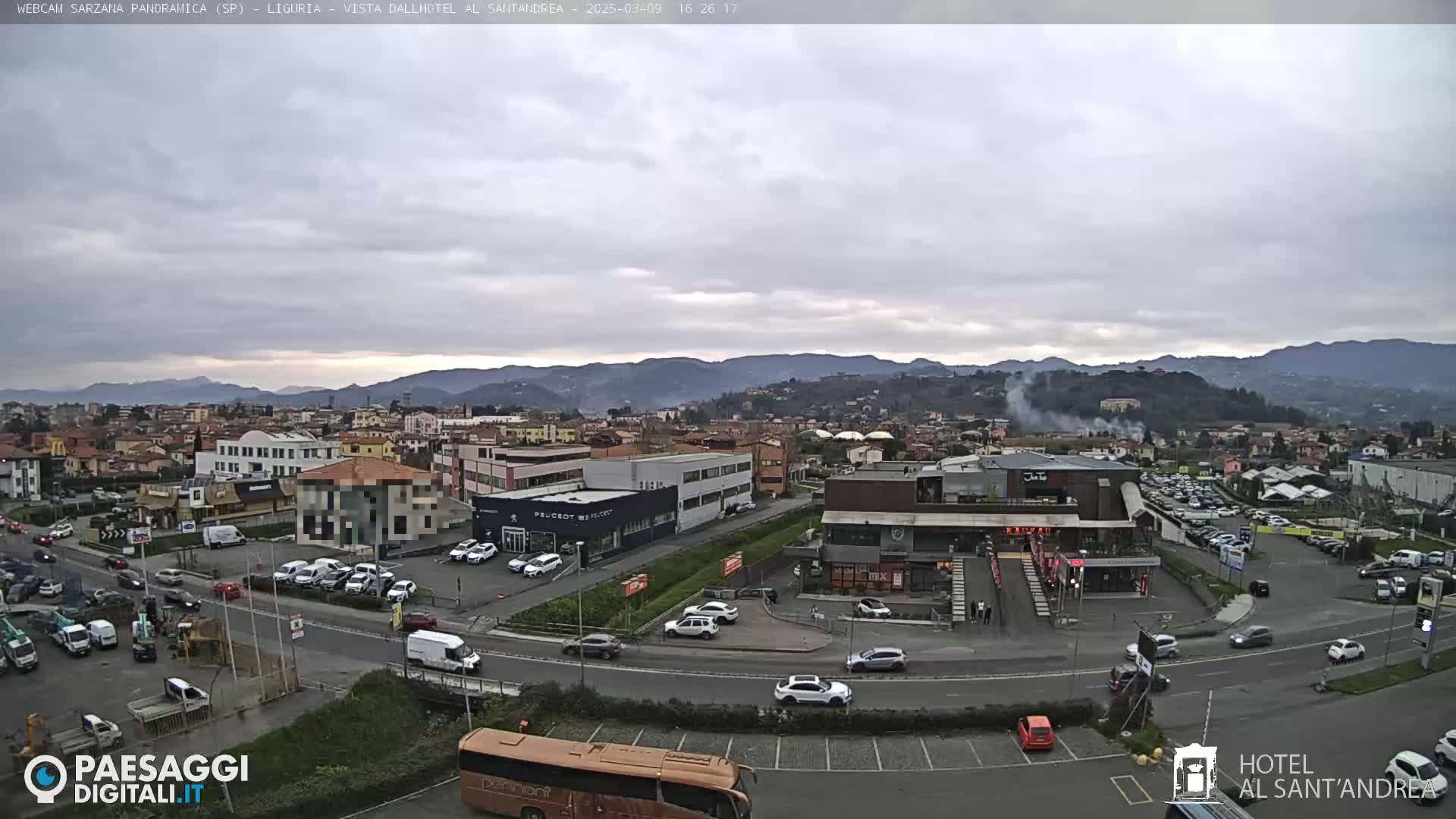 A sprawling town scene under an overcast sky, showing a mix of buildings, roads with moving vehicles, and a distant mountain range.