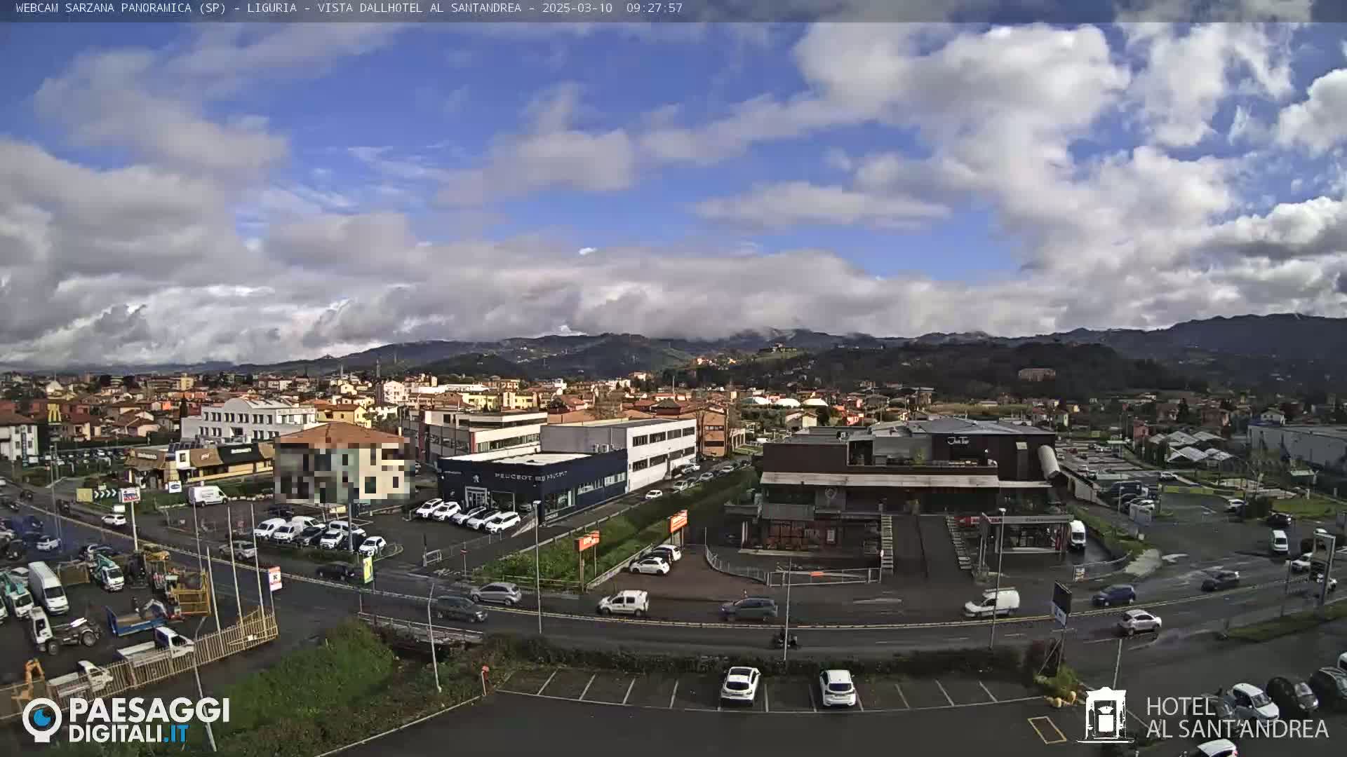 A partly cloudy day reveals a wide view of a town, featuring roads, buildings, and parked cars, with mountains visible in the background.