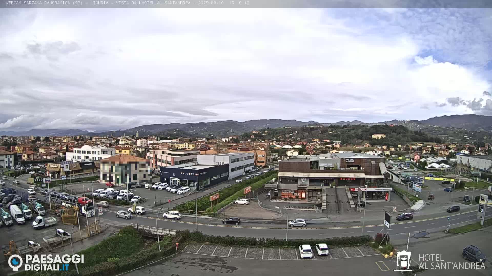 A partly cloudy day reveals a sprawling town nestled in a valley, surrounded by low, tree-covered mountains.