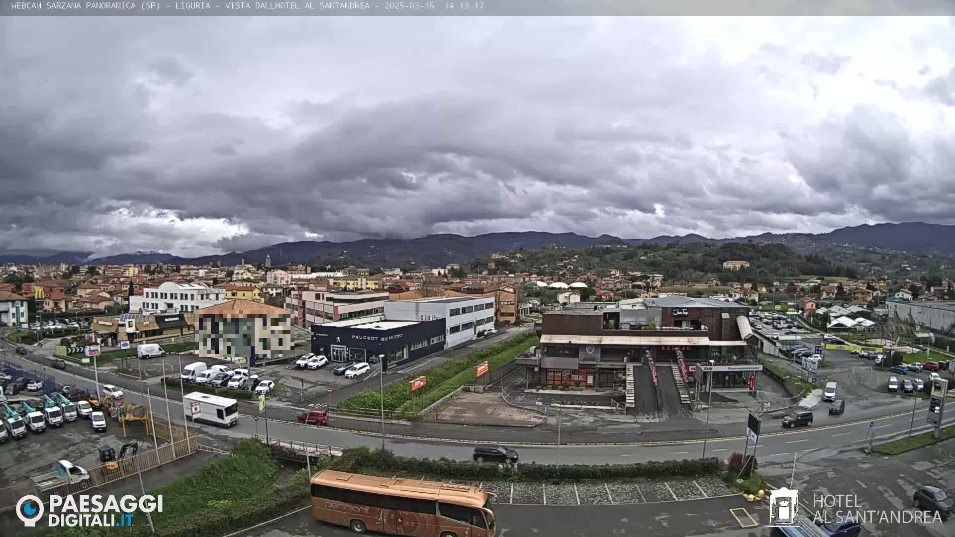 A wide shot of a town under a cloudy sky shows buildings, cars, and a bus in a parking lot, with mountains in the background.