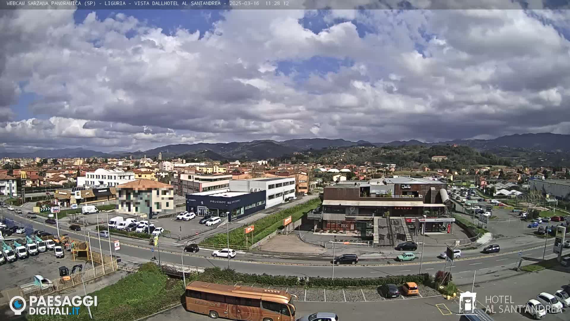 A partly cloudy day reveals a panoramic view of a town with buildings, roads, vehicles, and a bus, nestled amongst hills.