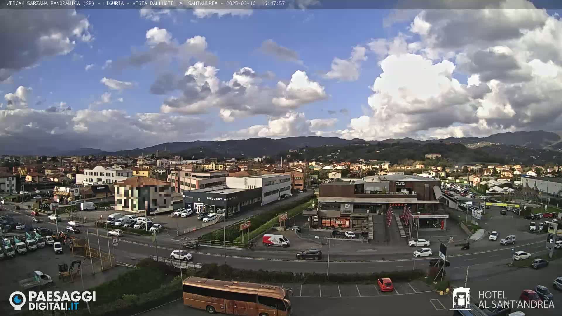 A partly cloudy day reveals a wide view of a town nestled against a mountain range, with numerous cars parked and driving along roads and in parking lots.