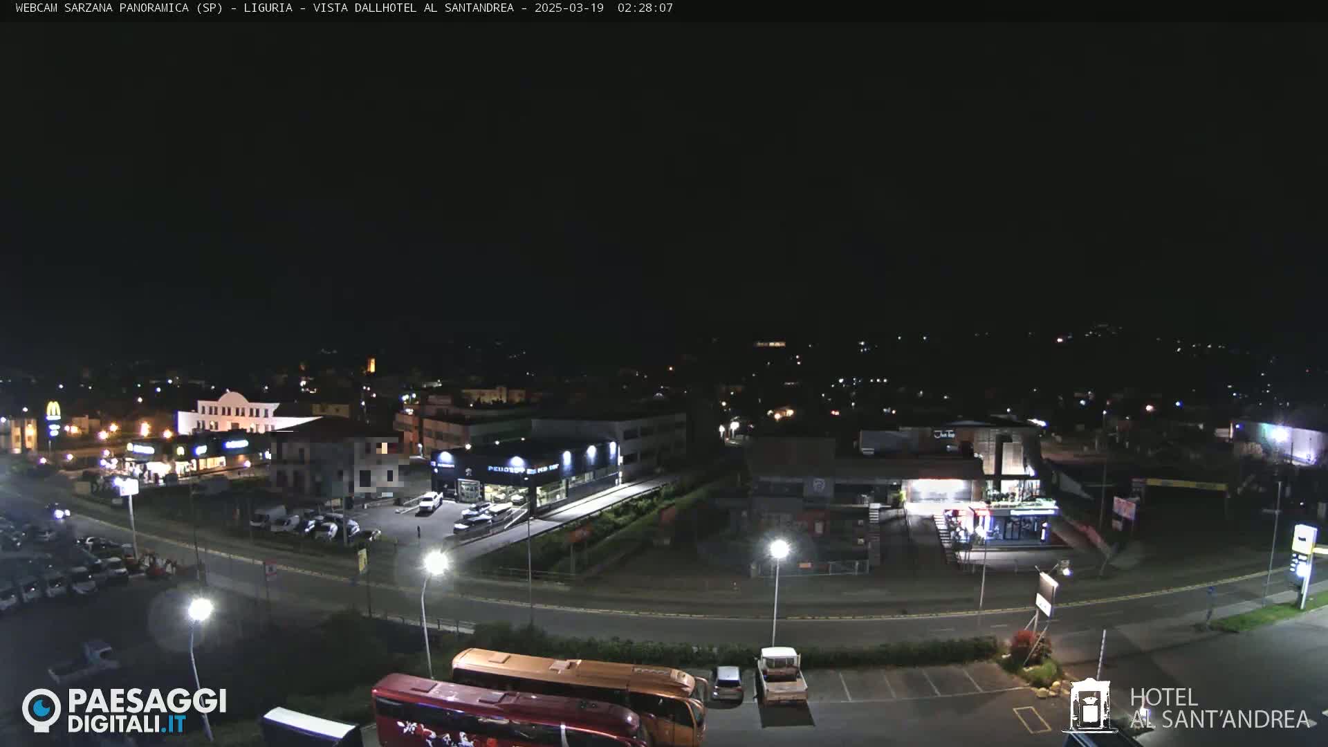 A nighttime aerial view of a town's outskirts, showing roads, buildings, parked cars, and buses, under a clear, dark sky.