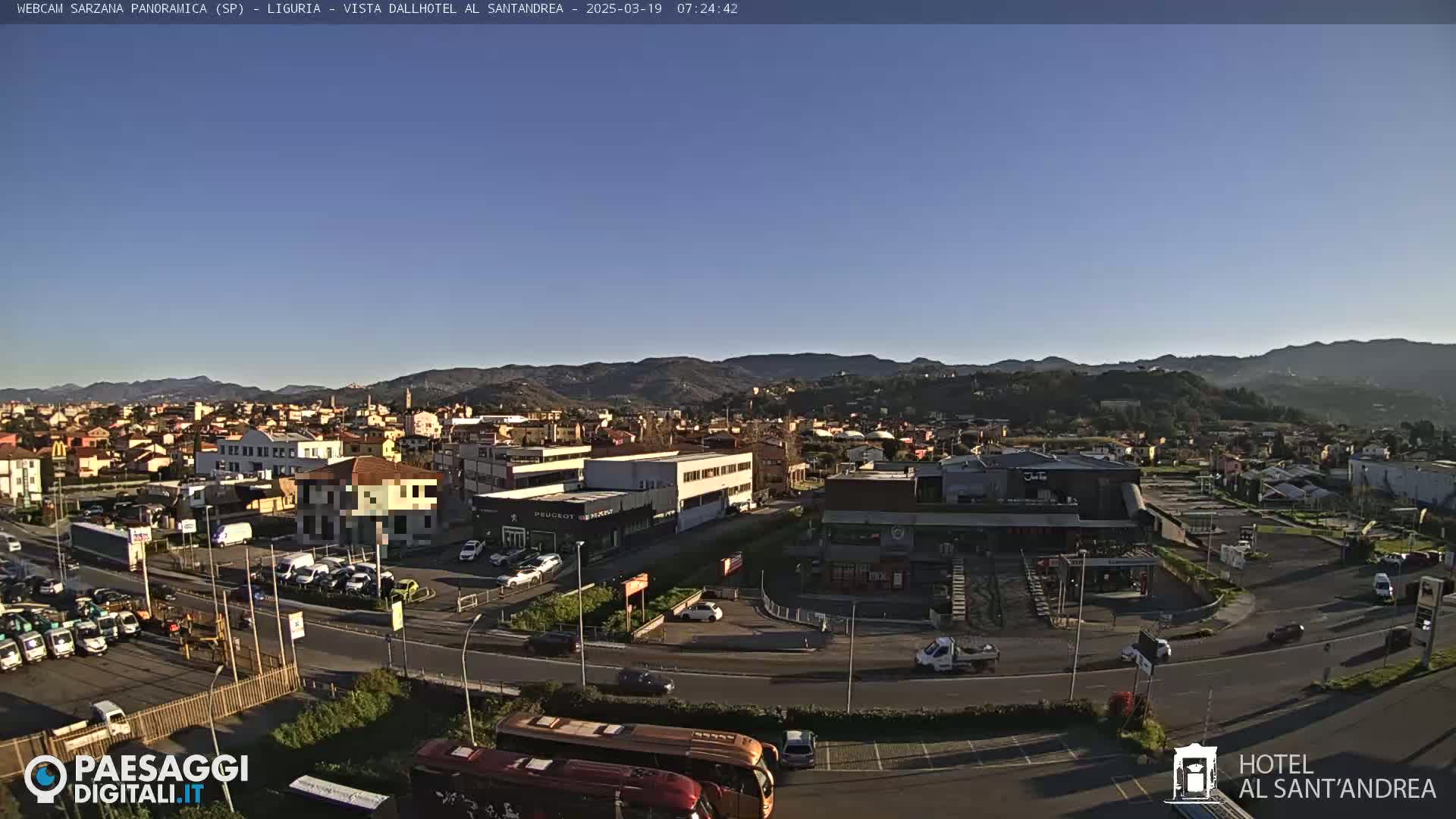 A clear, sunny day reveals a panoramic view of a town nestled in a valley, with mountains in the background and various vehicles parked in a lot near buildings.