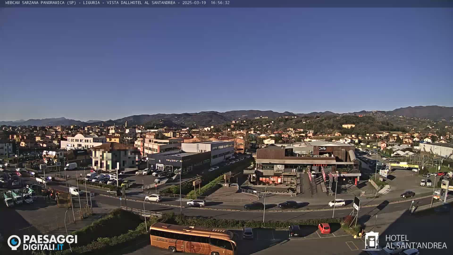 A sunny day reveals a panoramic view of a town nestled among hills, featuring a bustling road with cars and a large tour bus.