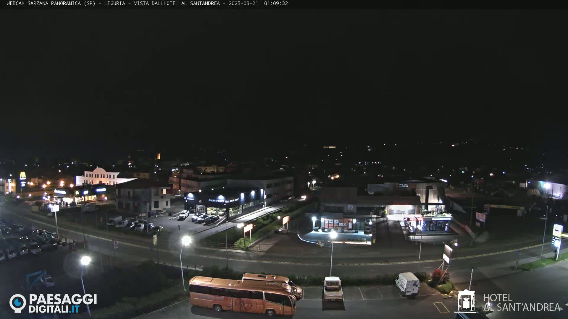 A nighttime, high-angle view of a town's outskirts showing illuminated buildings, roads, vehicles, and a large parking lot.