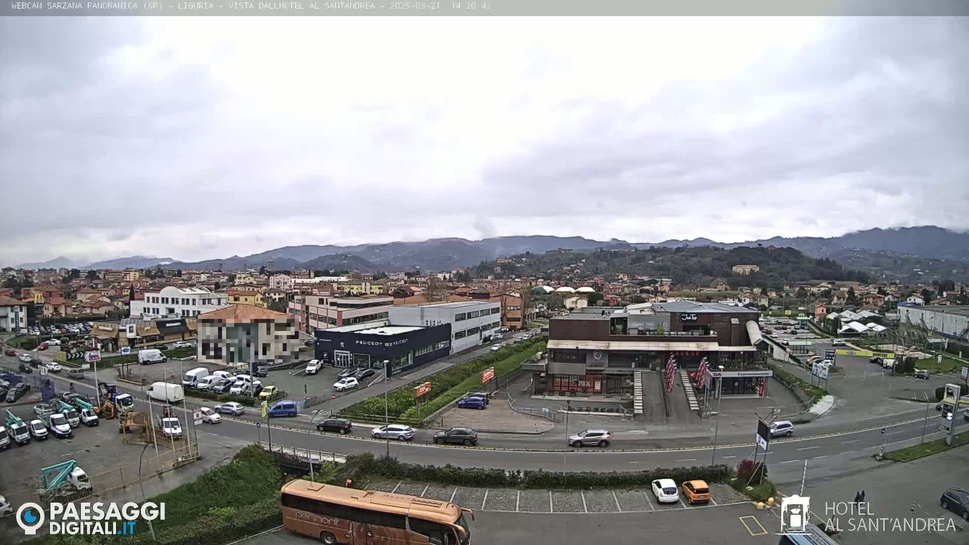A panoramic view of a town on an overcast day, showing roads, buildings, parked cars, and a bus, with mountains in the background.