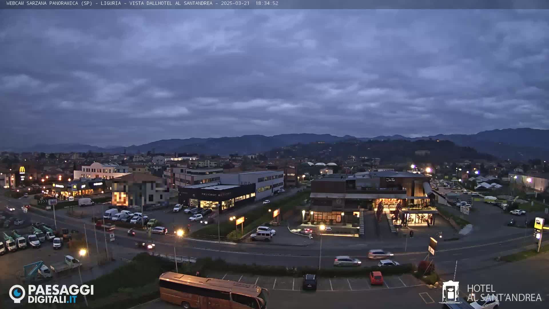 A twilight view of a town with buildings, roads, and parked cars under a cloudy sky.
