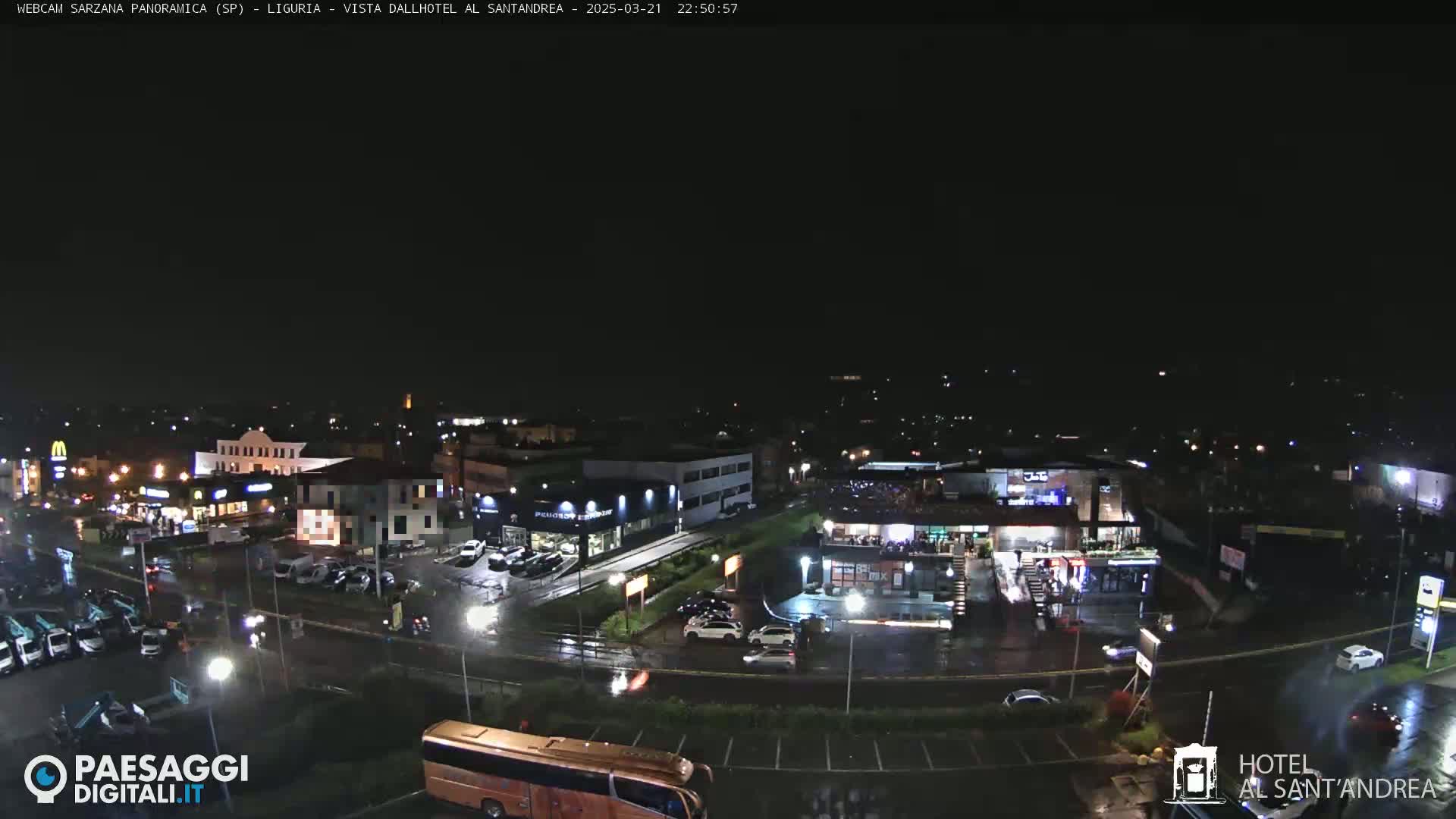A nighttime view of a wet parking lot and surrounding businesses, illuminated by streetlights and building lights.