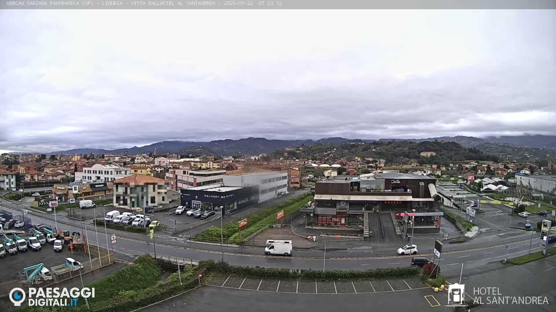 An overcast day reveals a sprawling town nestled in a valley, with mountains in the distance and various buildings, including a car dealership and a restaurant, visible at street level.
