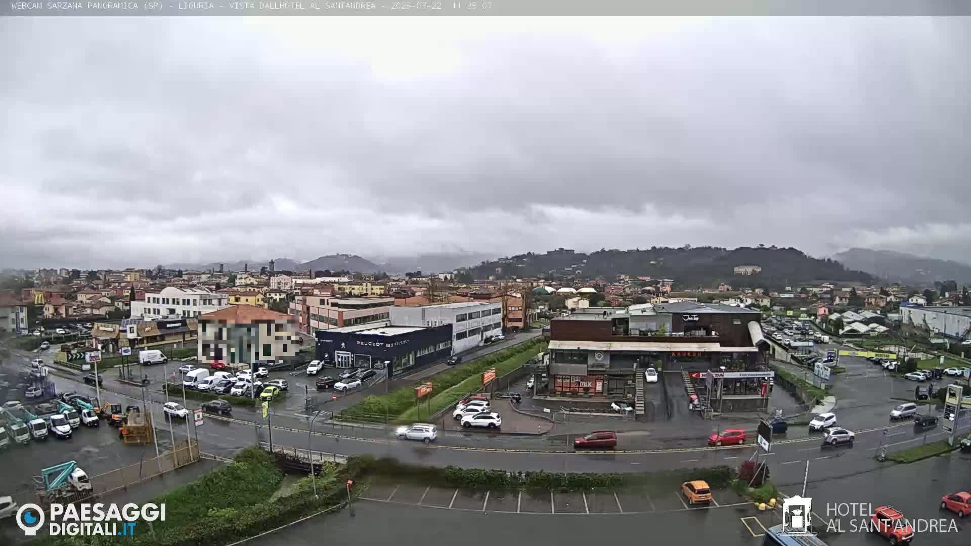 A wide shot of a town on a cloudy day shows wet roads and several cars parked in lots near various buildings.
