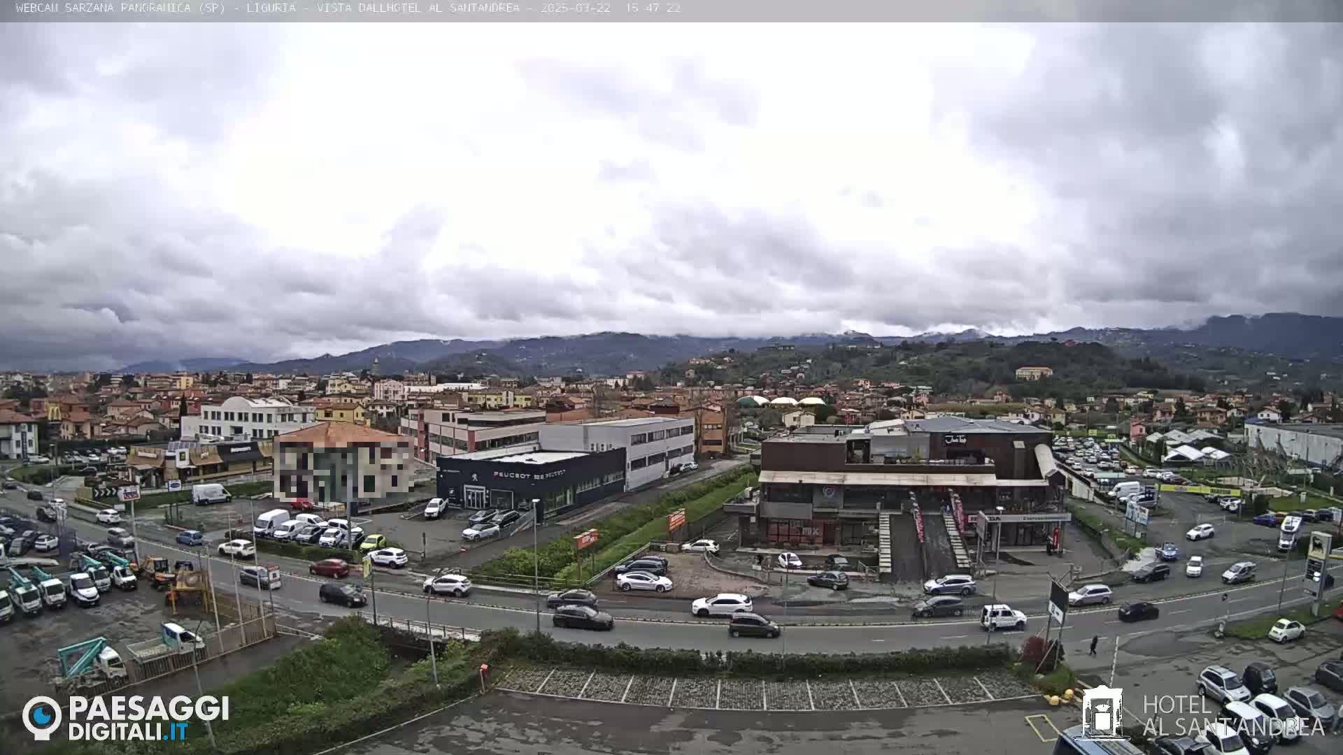 A cloudy day reveals a sprawling town with numerous buildings, cars parked in lots and driving on roads, and mountains in the background.