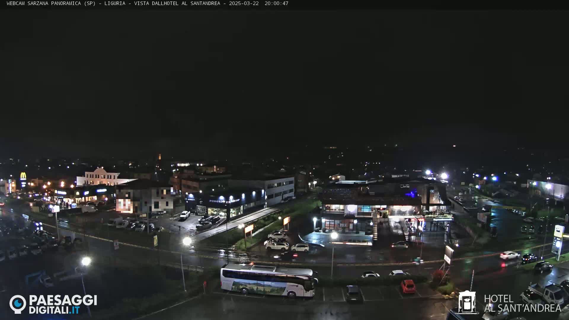 A nighttime aerial view of a town, showing roads, buildings, parked cars, and a bus, with wet pavement suggesting rain.