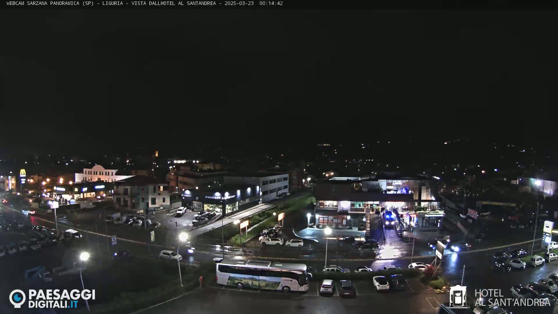 A nighttime, overhead view of a town’s street and parking lot with various buildings, cars, and a bus, under slightly wet conditions.