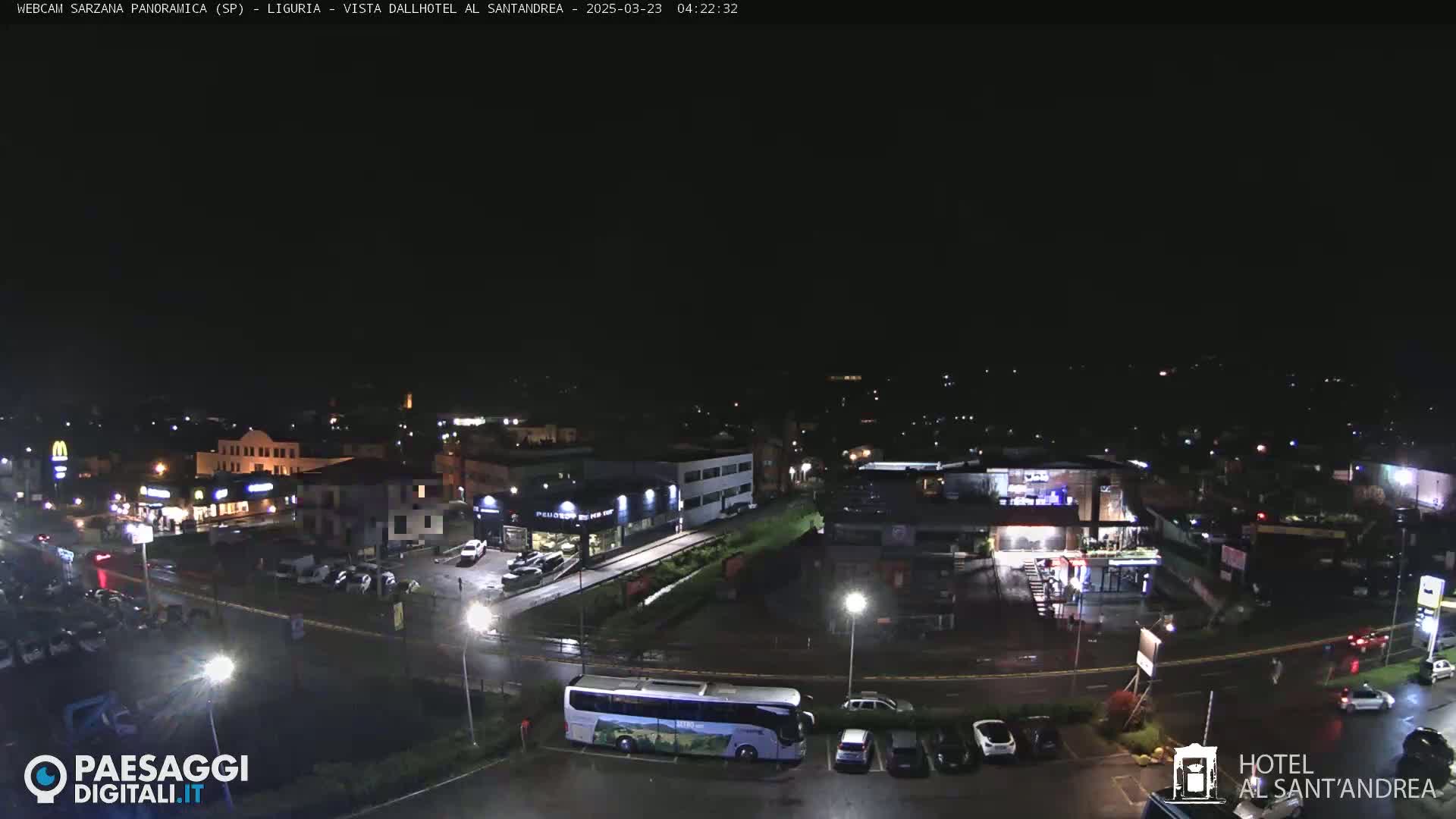 A nighttime, wet view of a town with buildings, roads, vehicles, and a large bus in a parking lot.