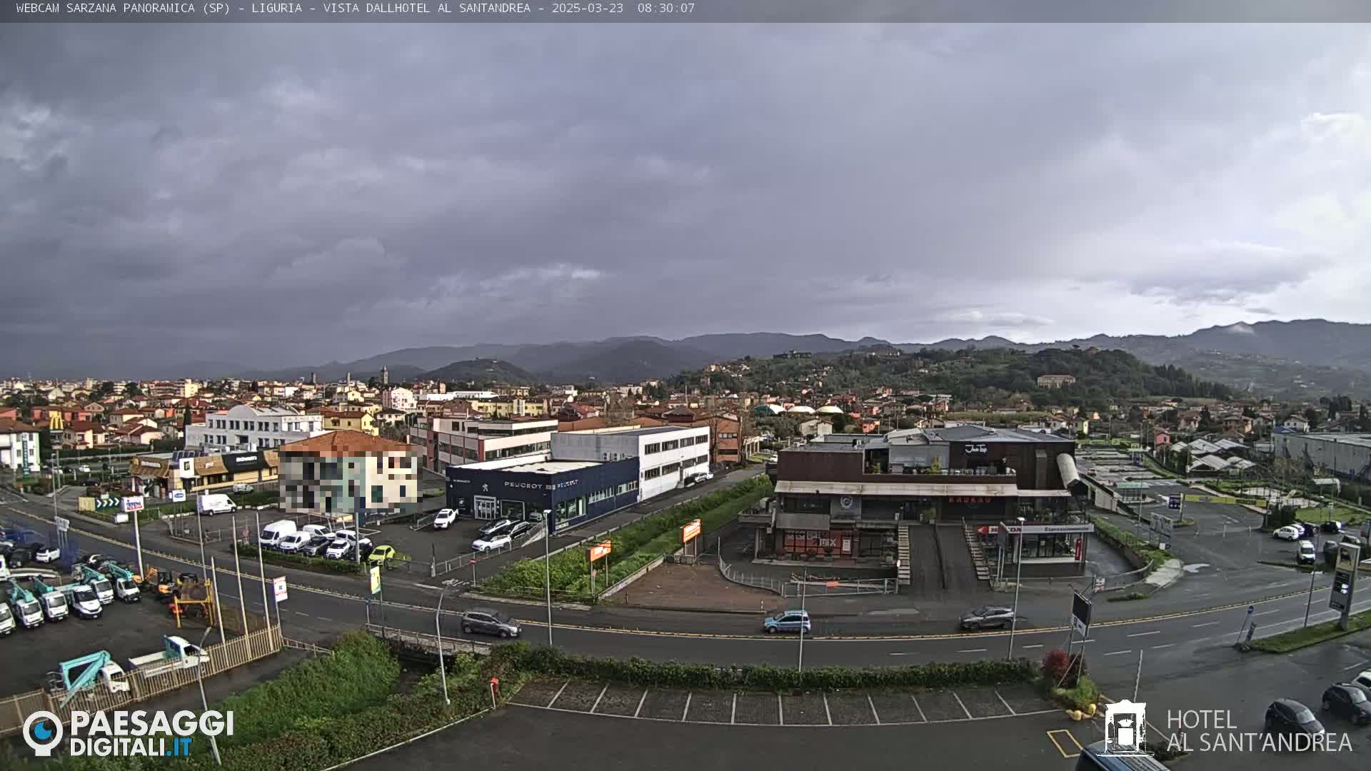 A panoramic view of a town under a cloudy sky, showing buildings, roads, cars, and distant mountains.