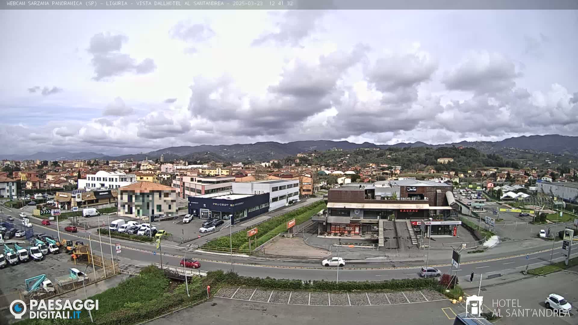 A partly cloudy day reveals a sprawling town nestled against a backdrop of rolling hills, with various buildings, vehicles, and a parking lot visible in the foreground.