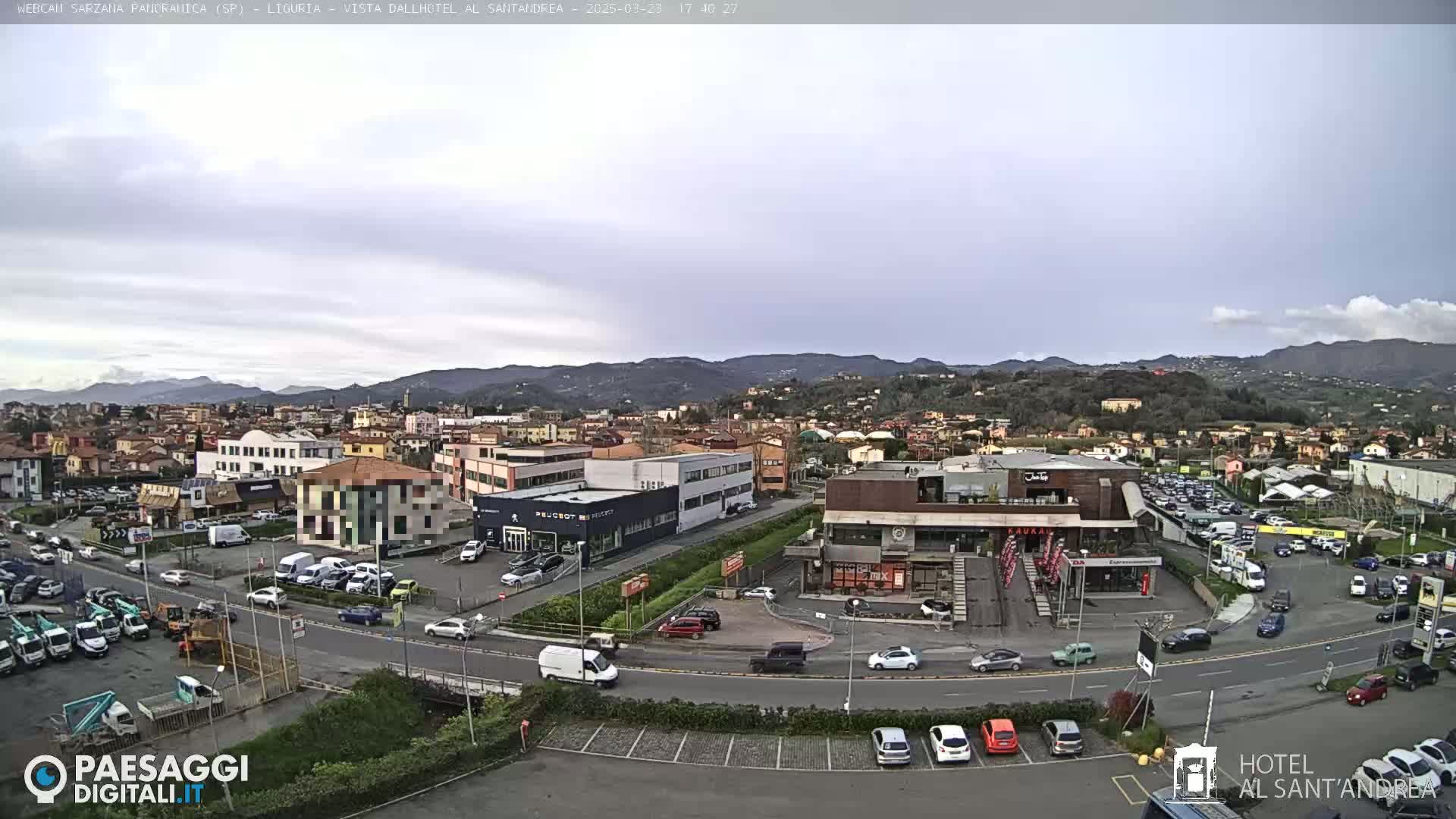 A wide shot of a town situated in a valley, under a cloudy sky, shows roads with cars and buildings, including businesses and parking lots.