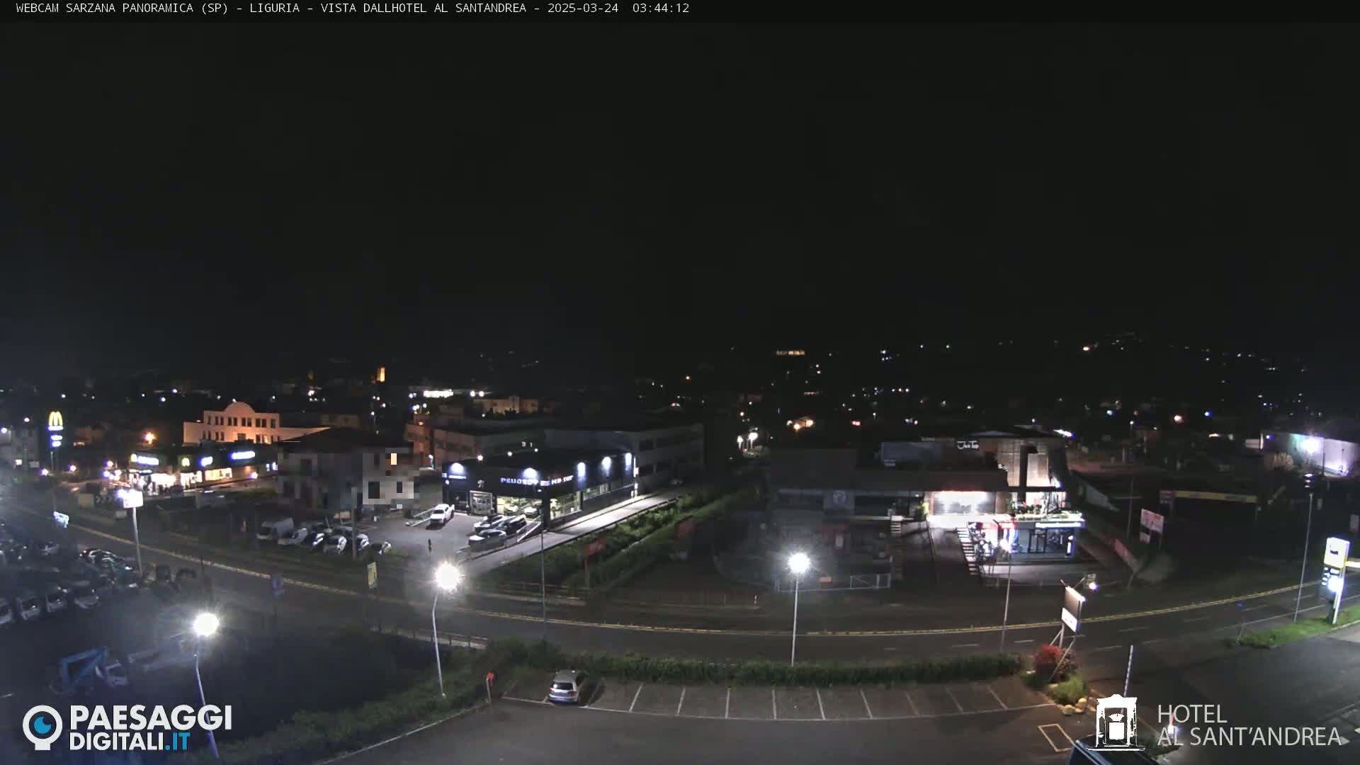 A nighttime, high-angle view of a town's outskirts, showing roads, buildings, and sparsely parked cars under a clear, dark sky.