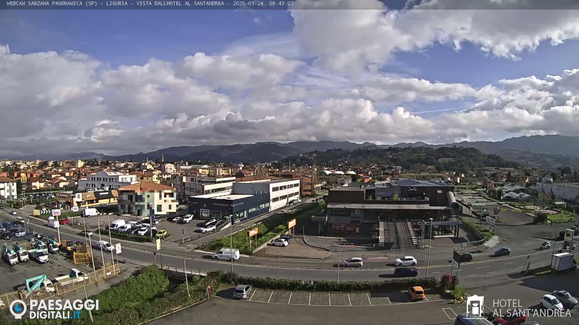 A partly cloudy day reveals a panoramic view of a town nestled in a valley, surrounded by mountains, with cars parked in lots and along roads.