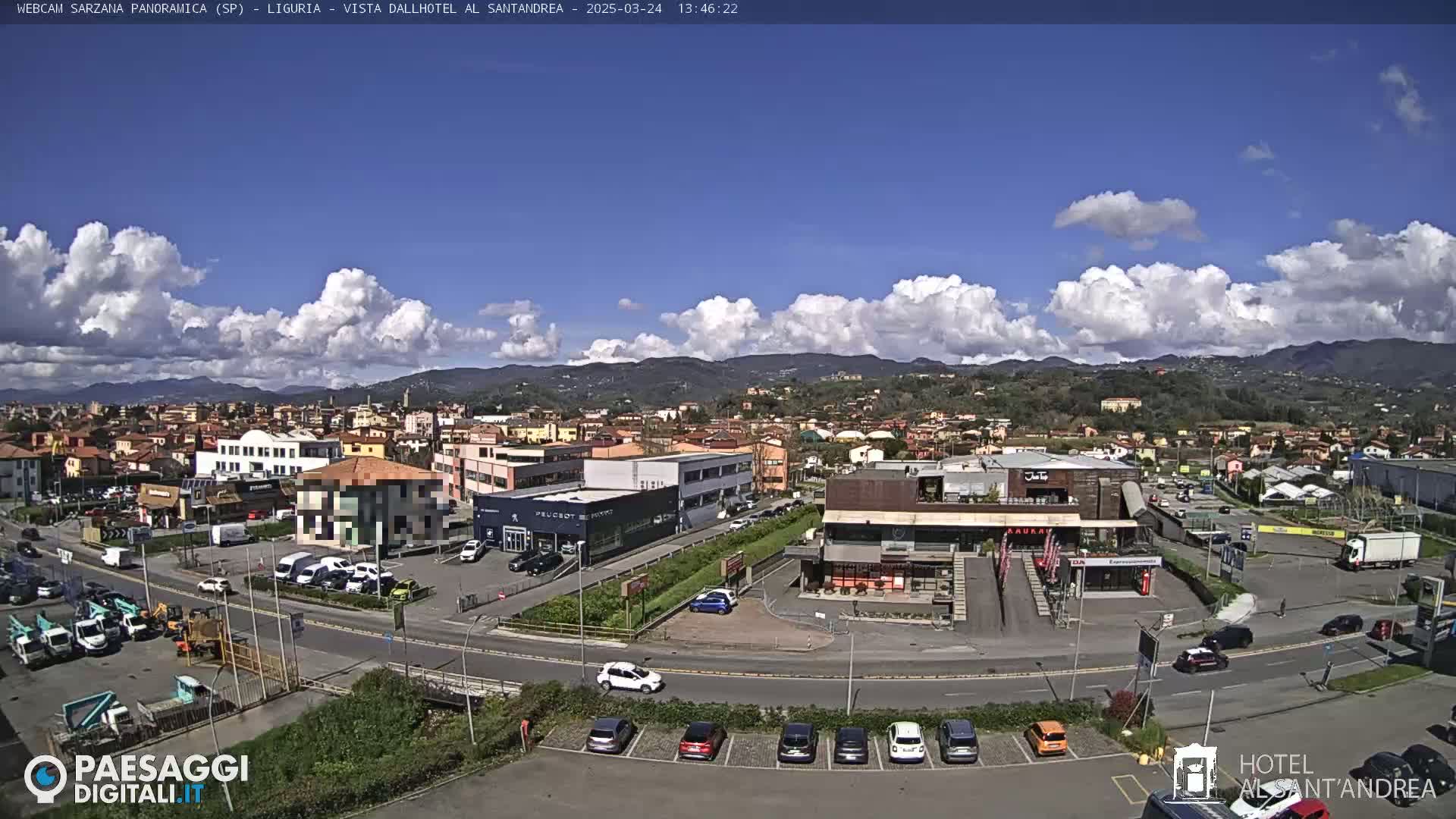 A sunny day with scattered cumulus clouds overlooks a town with various buildings, including a car dealership and a restaurant, and a parking lot with several cars.
